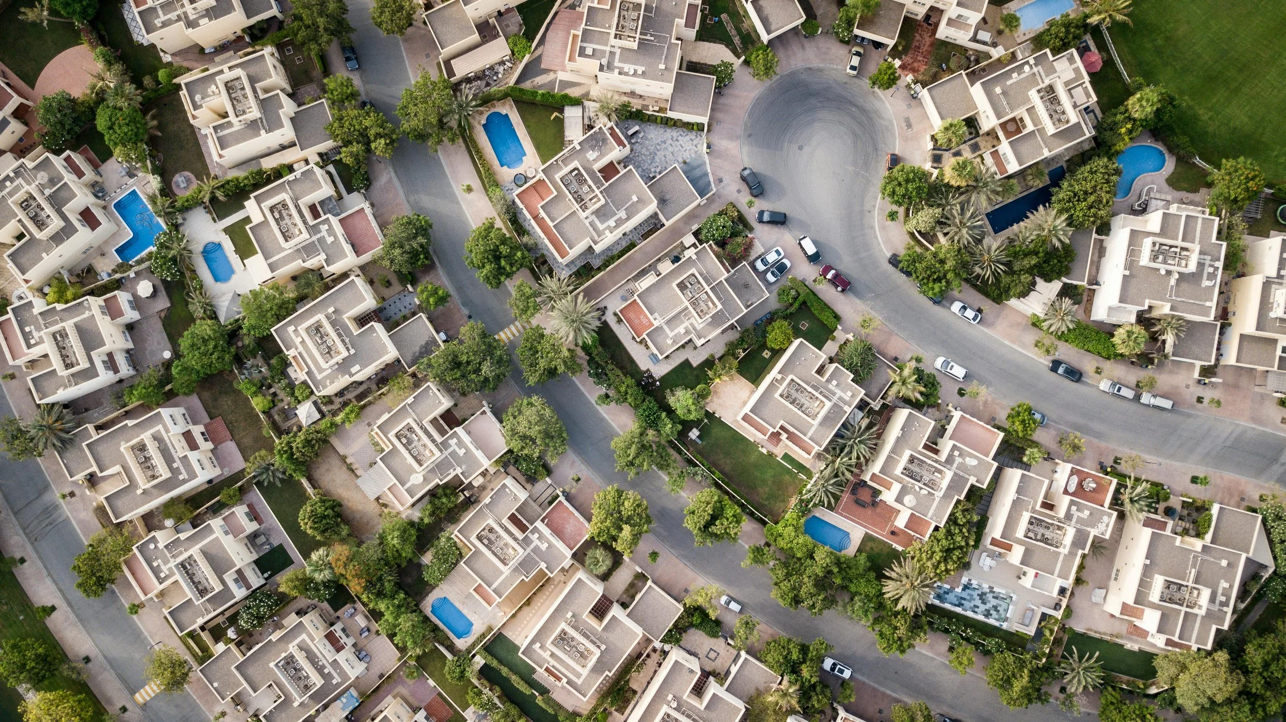 Imagem aérea de um condomínio com casas com quintais, piscinas pequenas, árvores e ruas pavimentadas, incluindo um círculo de rua no centro.