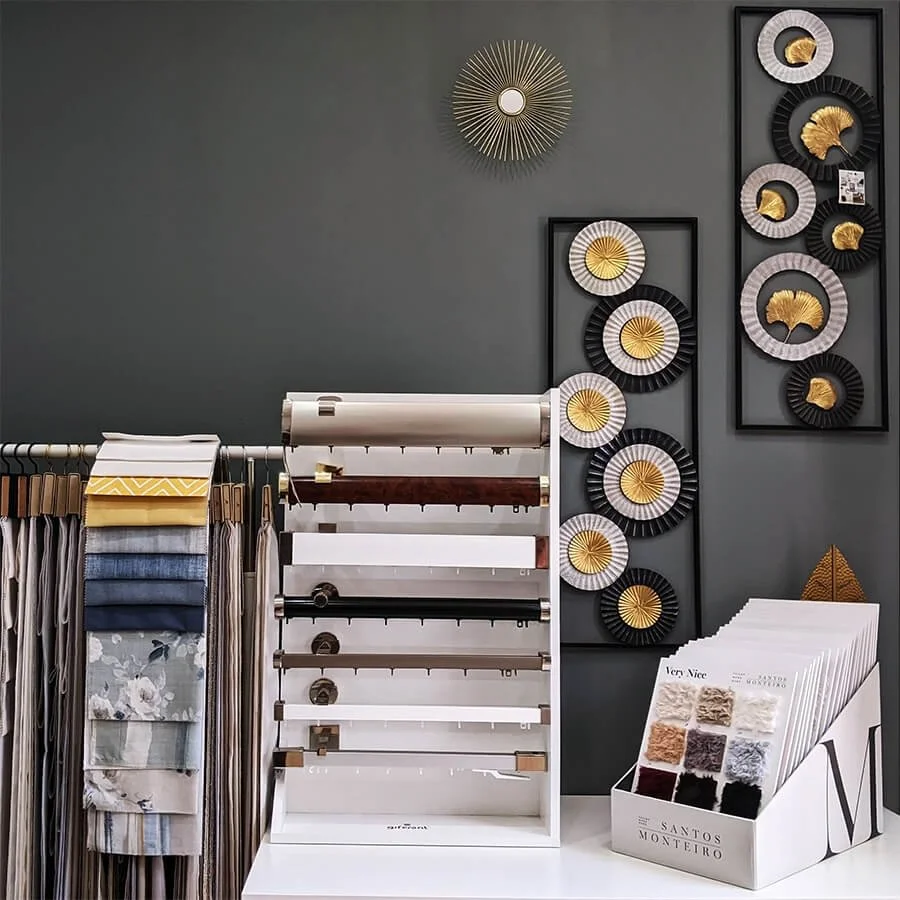 Mostruário de tecidos e papéis decorativos com parede de fundo cinza escuro, decorada com vários quadros de peças decorativas em preto, branco e detalhes dourados.