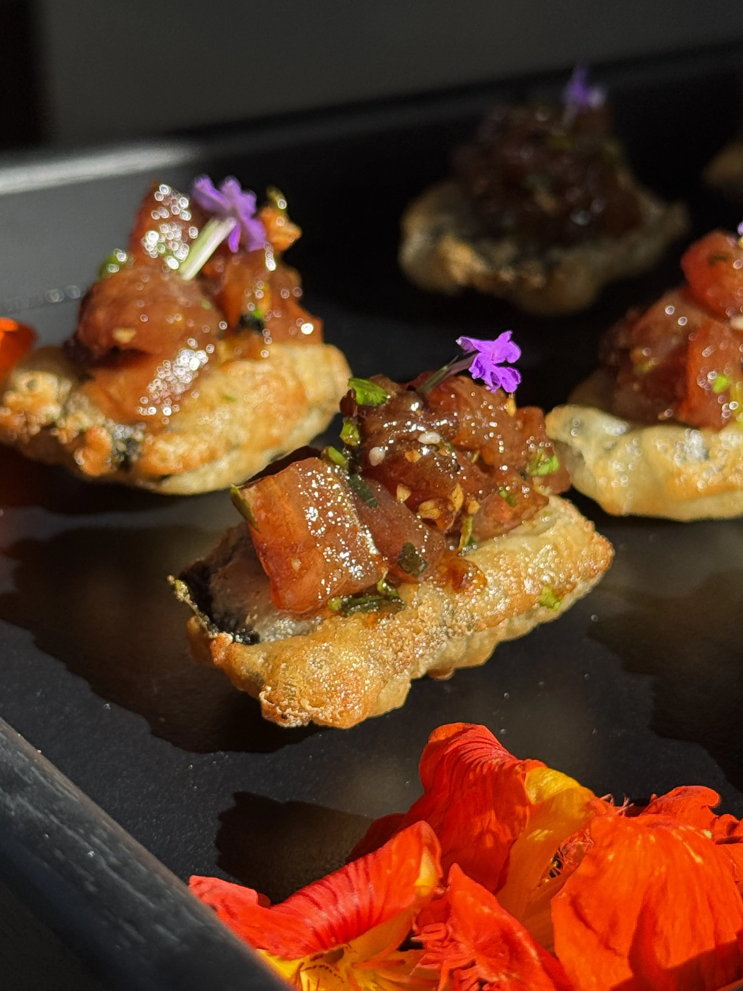 Plato con carne en salsa, decorada con flores comestibles moradas, servido con vegetales y salsas coloridas