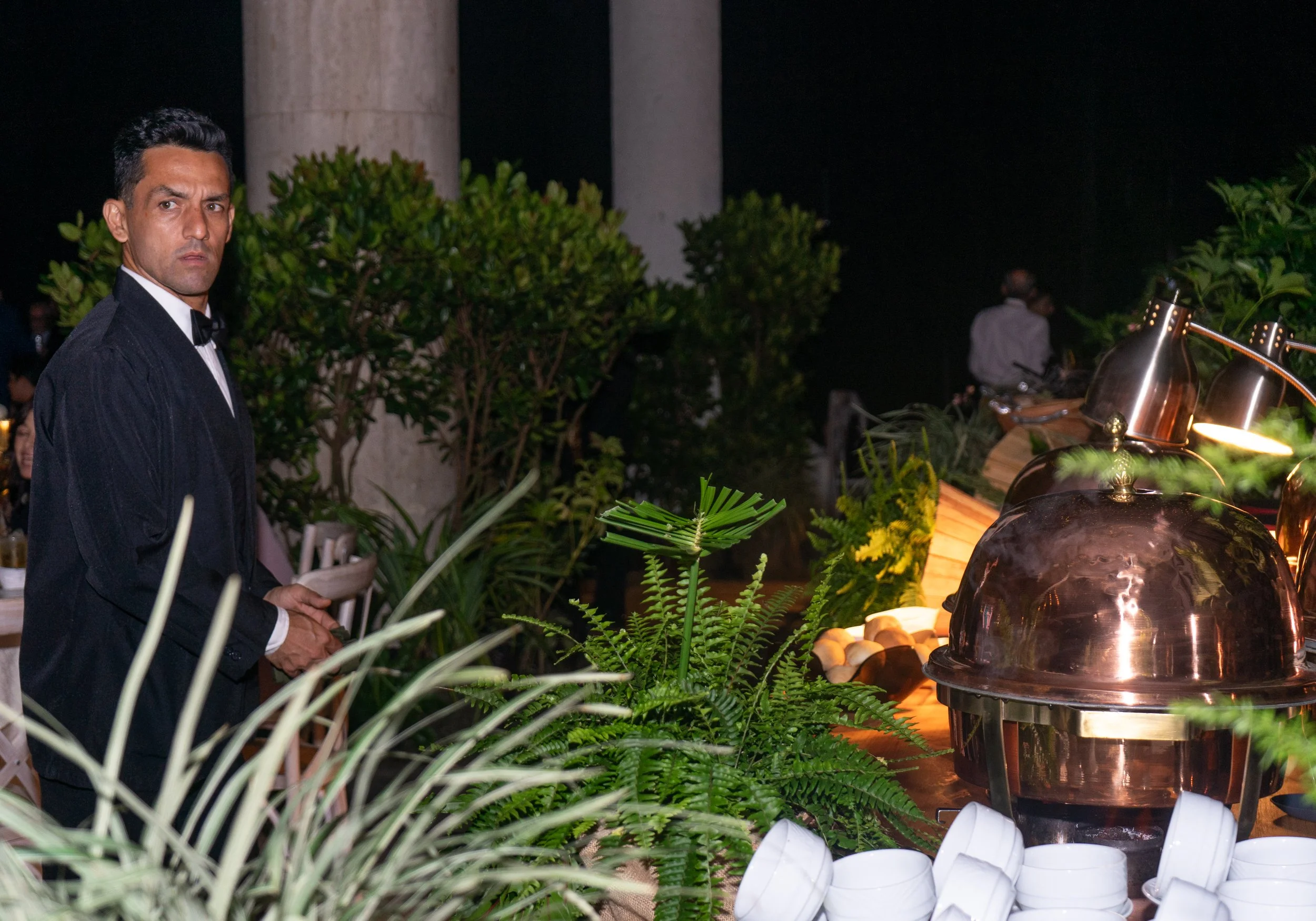 Hombre en traje y moño en una boda o evento formal, en un ambiente decorado con plantas, fuentes de agua y vajilla.