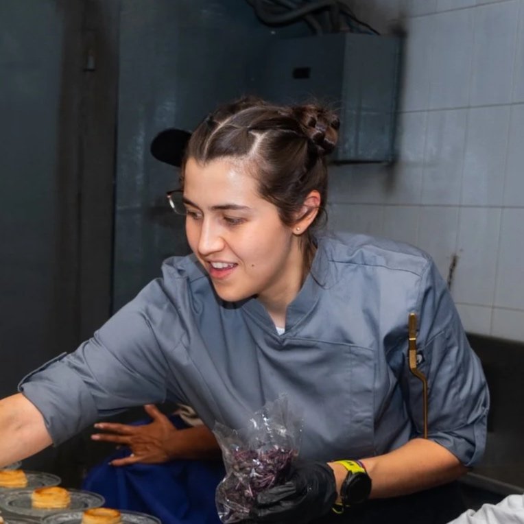 Una mujer con uniforme de chef decorando o colocando algo en unos pequeños postres en una cocina profesional.