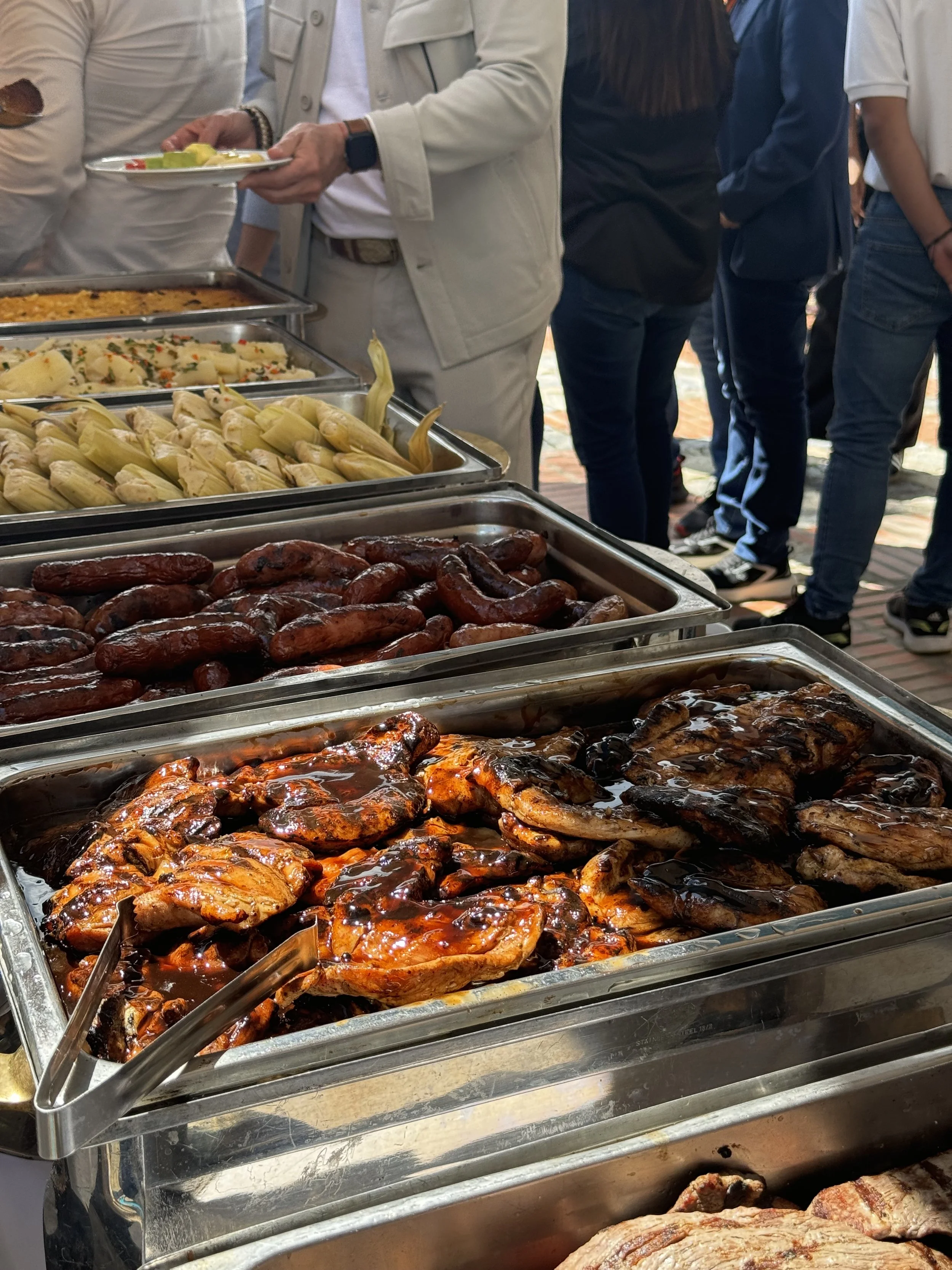 Variedad de alimentos en una mesa de buffet, incluyendo salchichas, pollo en salsa, tamales, papas y otros platillos, con personas sirviéndose comida.