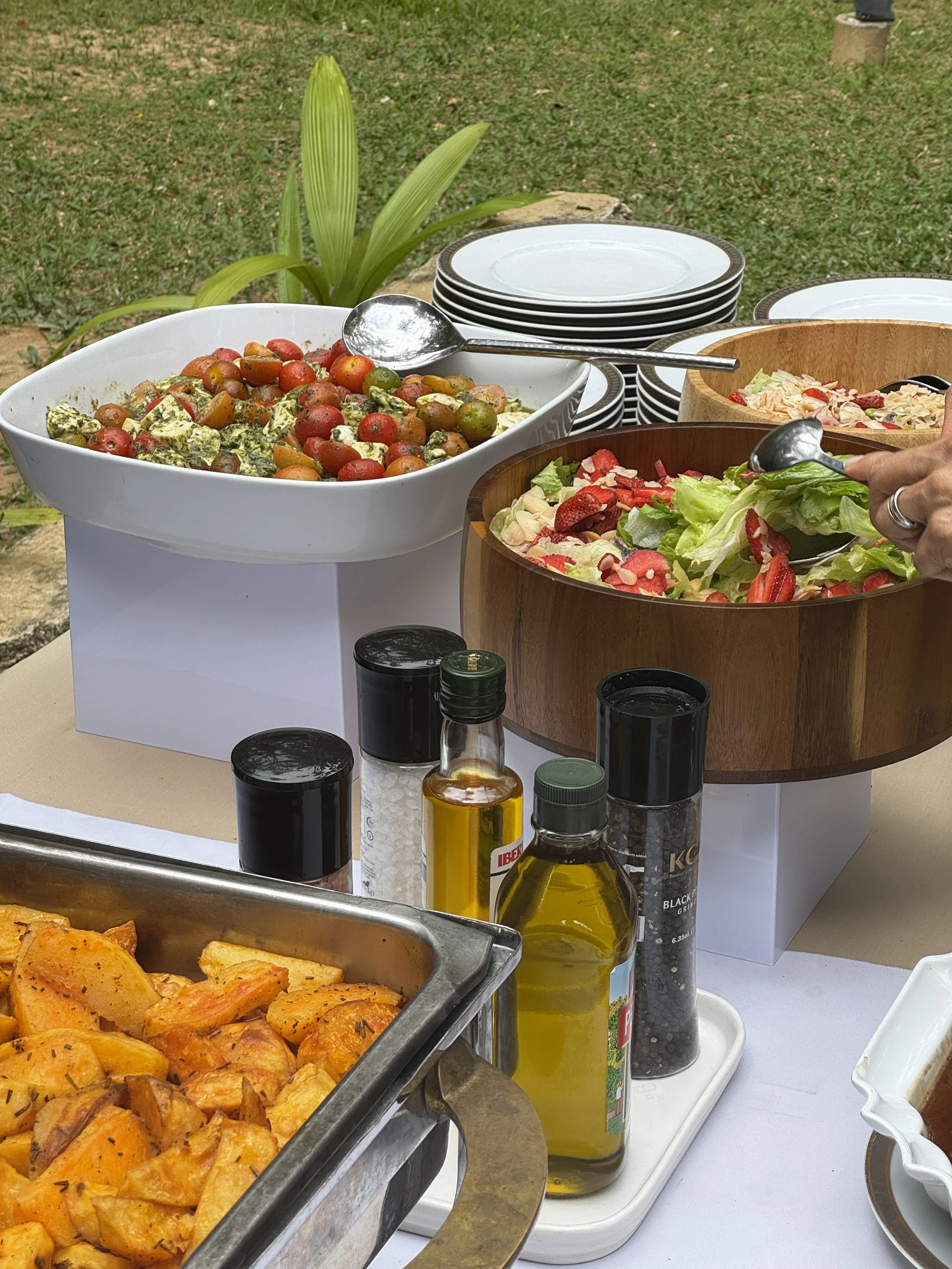 En la mesa hay ensaladas con jitomates cherry y fresa, un plato con papas a la francesa, varias condimentos y aceites, y platos desocupados en el fondo.