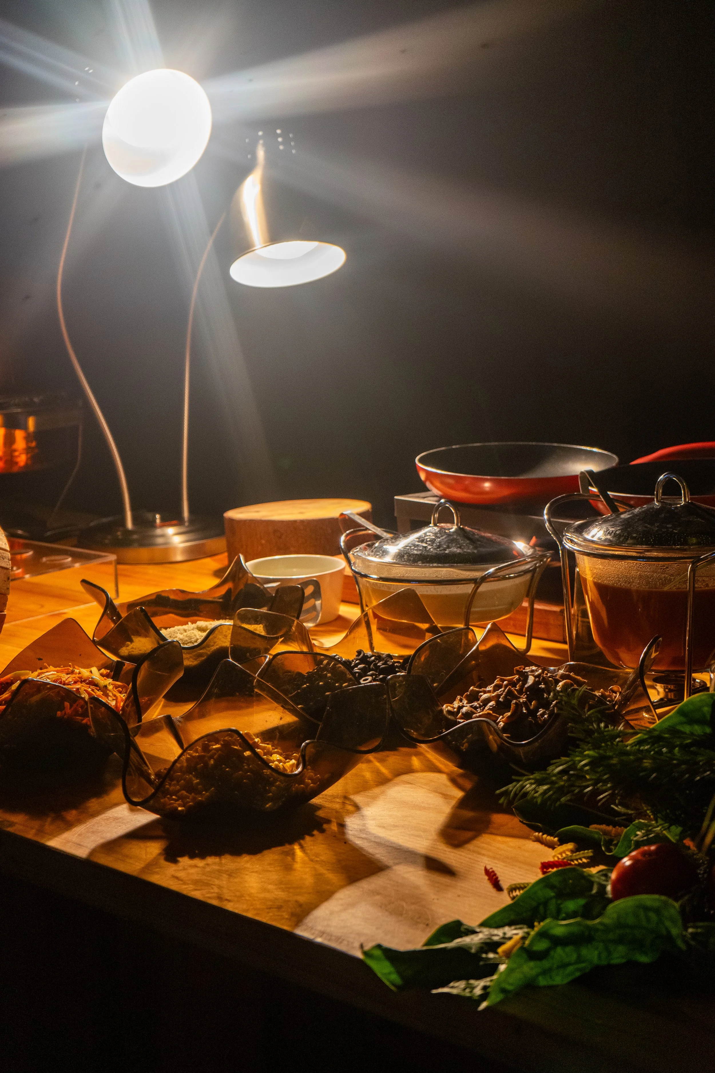Vista de una mesa con variedad de platillos y ingredientes, iluminada por lámparas de luz cálida en un ambiente de cocina o buffet.