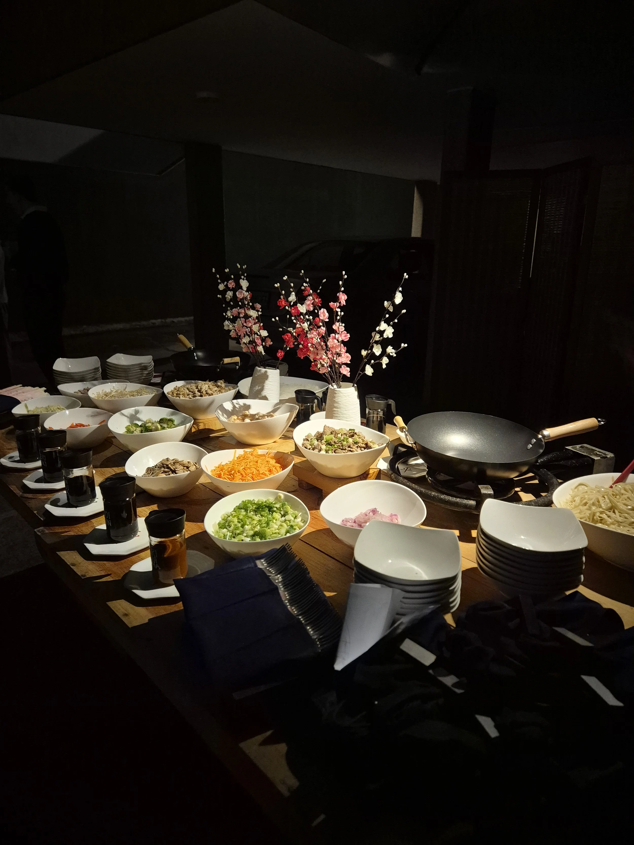 Mesa con diferentes platillos y comida, adornada con flores de cerezo en un jarrón, en un ambiente oscuro.
