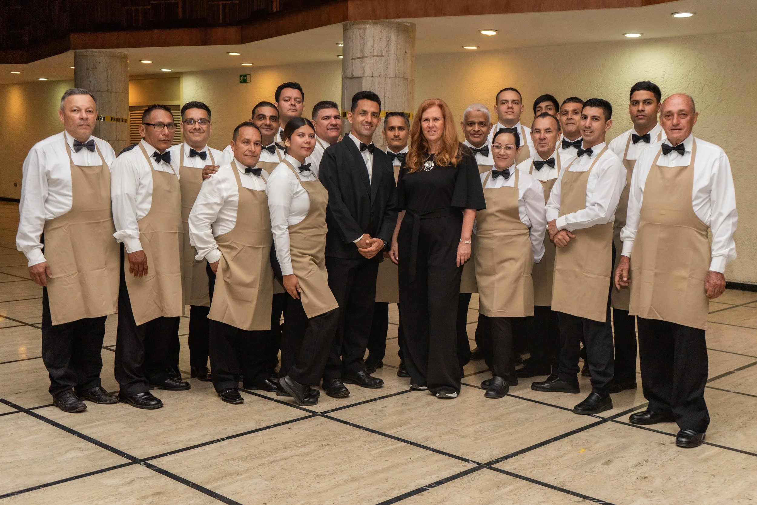 Grupo de personas en uniforme de camareros, con hombres y mujeres, algunos con delantales beige y otros con trajes negros con corbatas, en interior de edificio, posiblemente en evento o restaurante, con paredes de color crema y columnas de concreto visible.