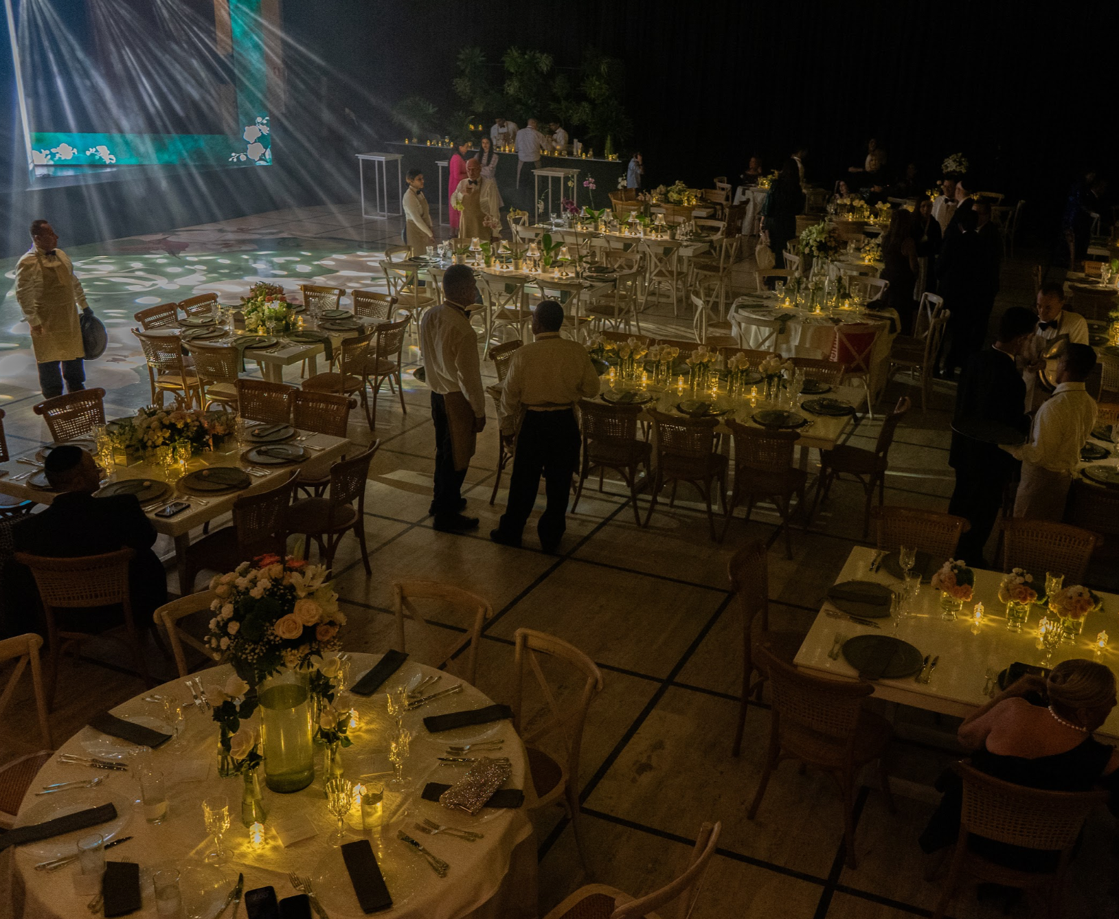Una sala de banquetes decorada con mesas y sillas, con varias personas y camareros, iluminación suave y centros de mesa con flores, en un evento formal por la noche.
