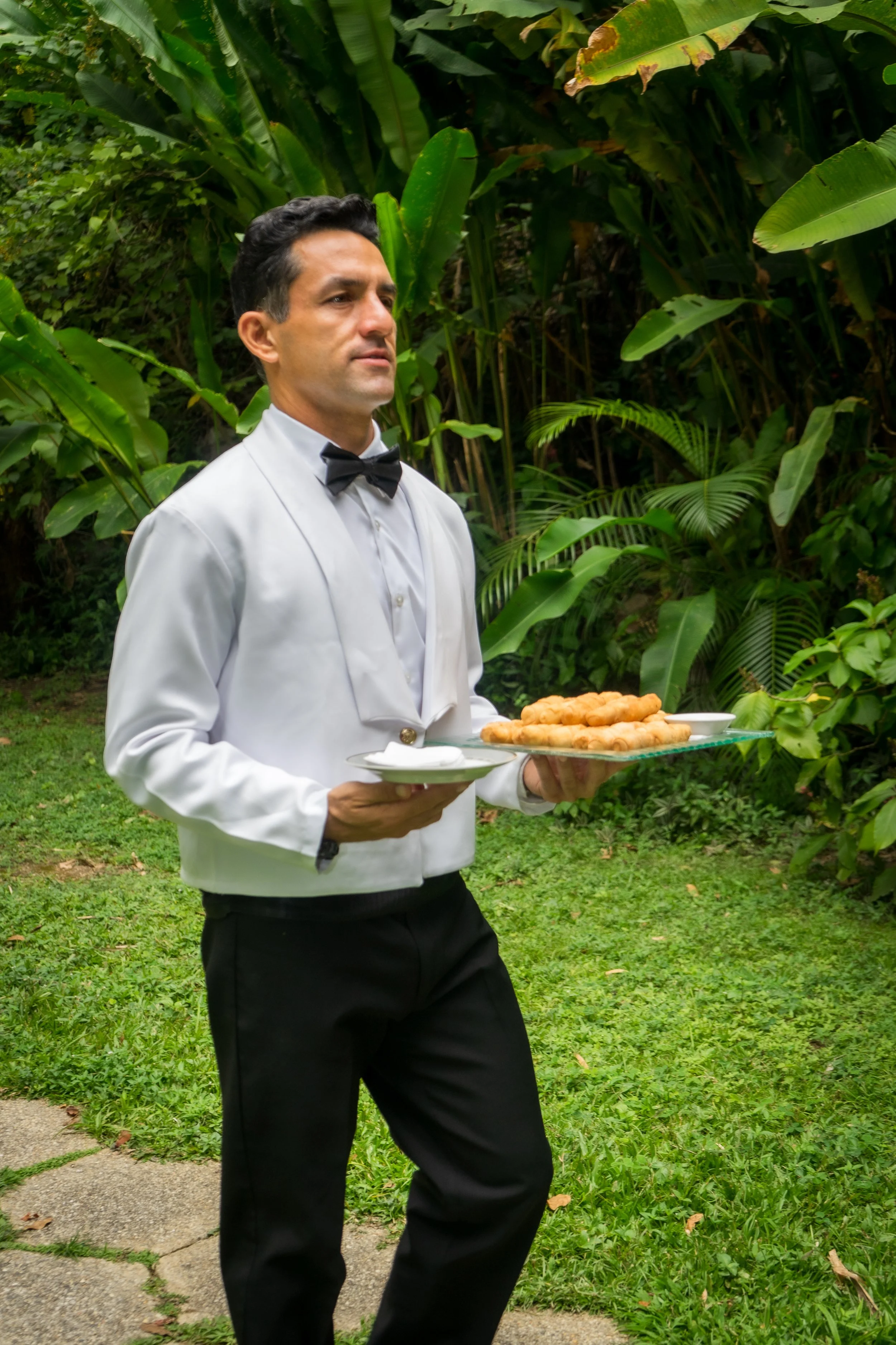 Un mayordomo con uniforme formal de camisa blanca y pajarita negra sostiene una bandeja con comida en un entorno natural con muchas plantas verdes.