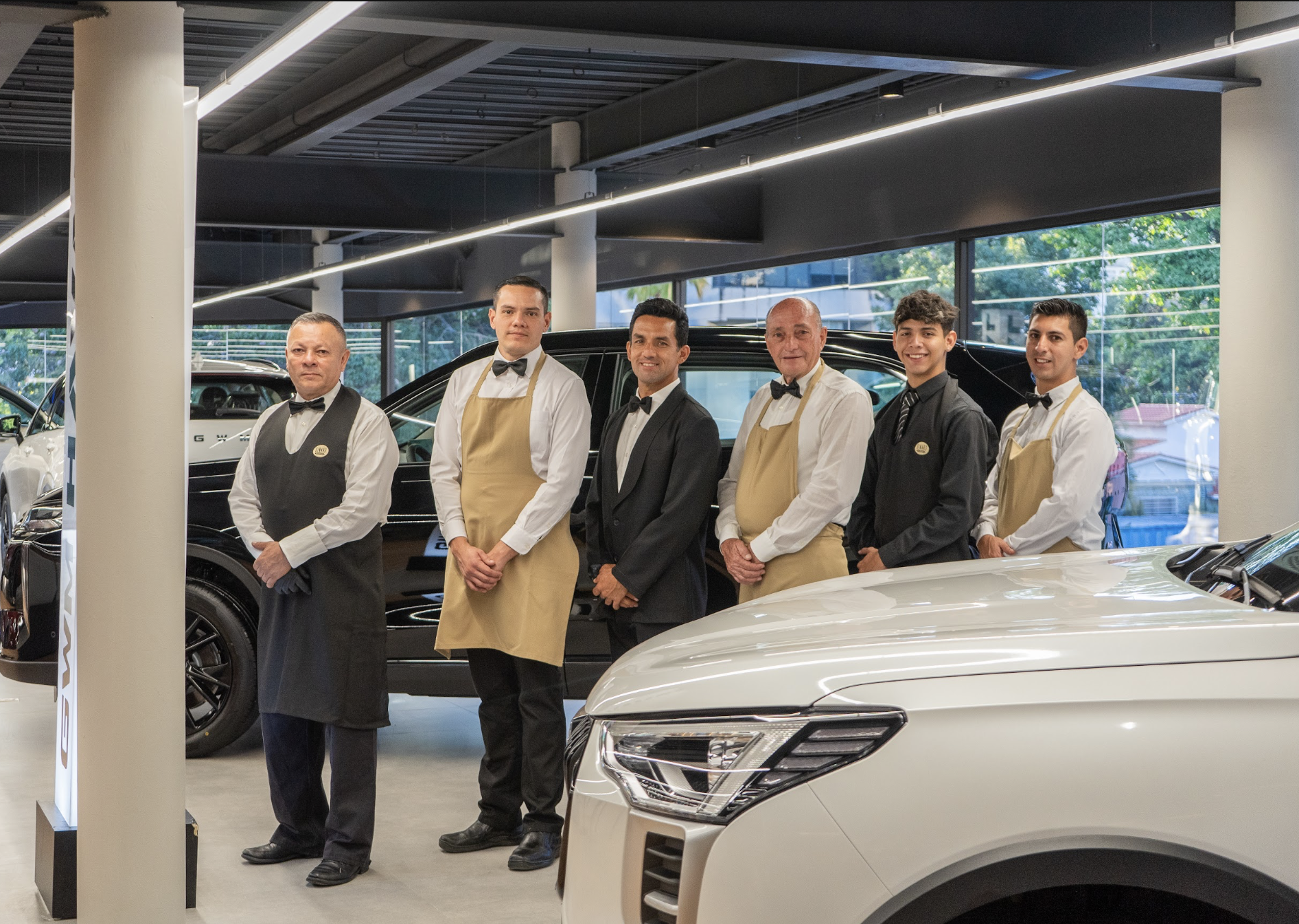 Grupo de seis empleados de un concesionario de autos, algunos con delantales y todos con corbata o pajarita, parados delante de varios autos de lujo en un showroom.