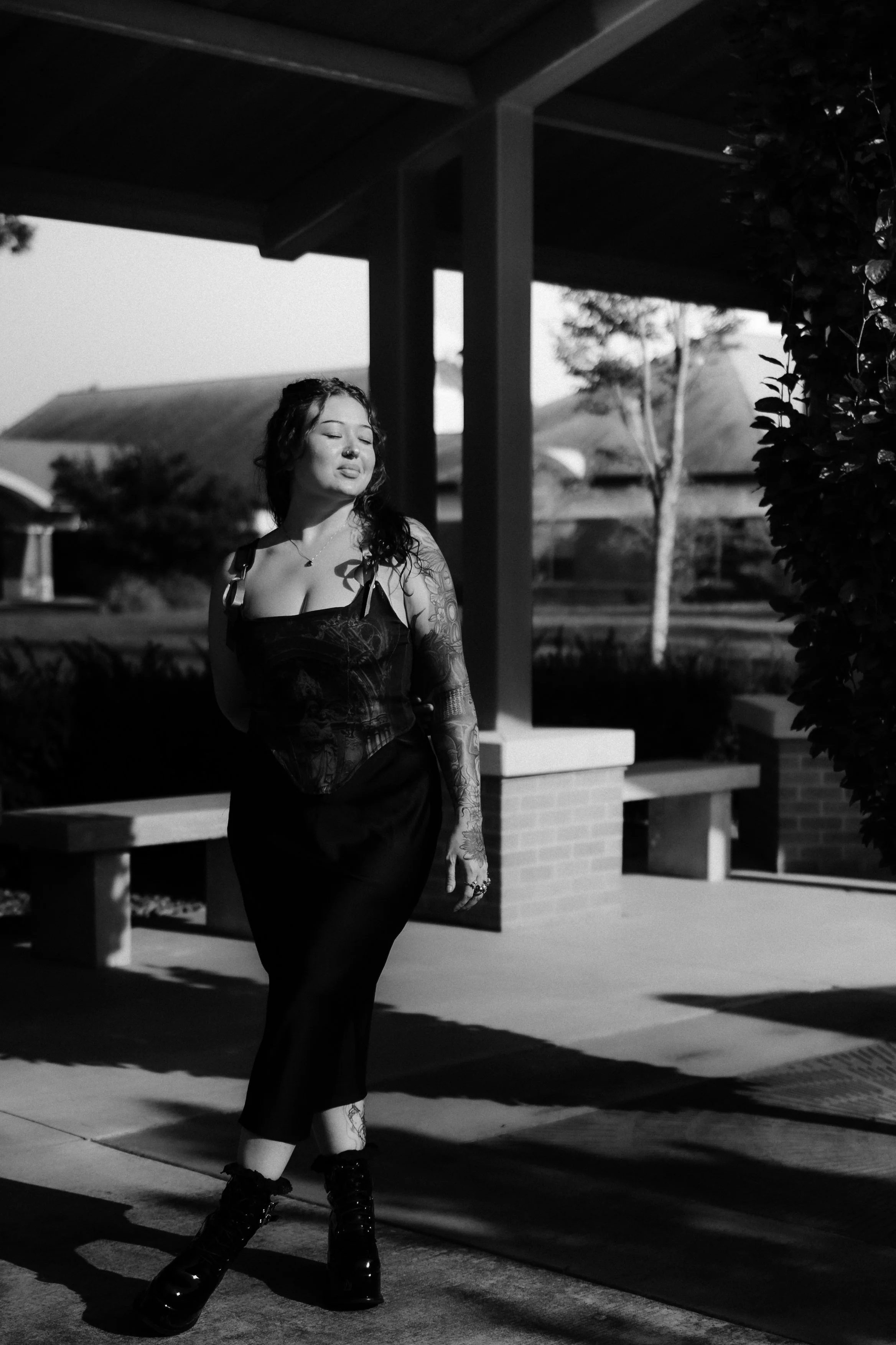 A woman with tattoos on her arm and leg is walking outdoors at night. She is wearing a strapless top, long pants, and chunky boots. She has curly hair and is putting her tongue out slightly. The background shows a building, trees, and a bench.