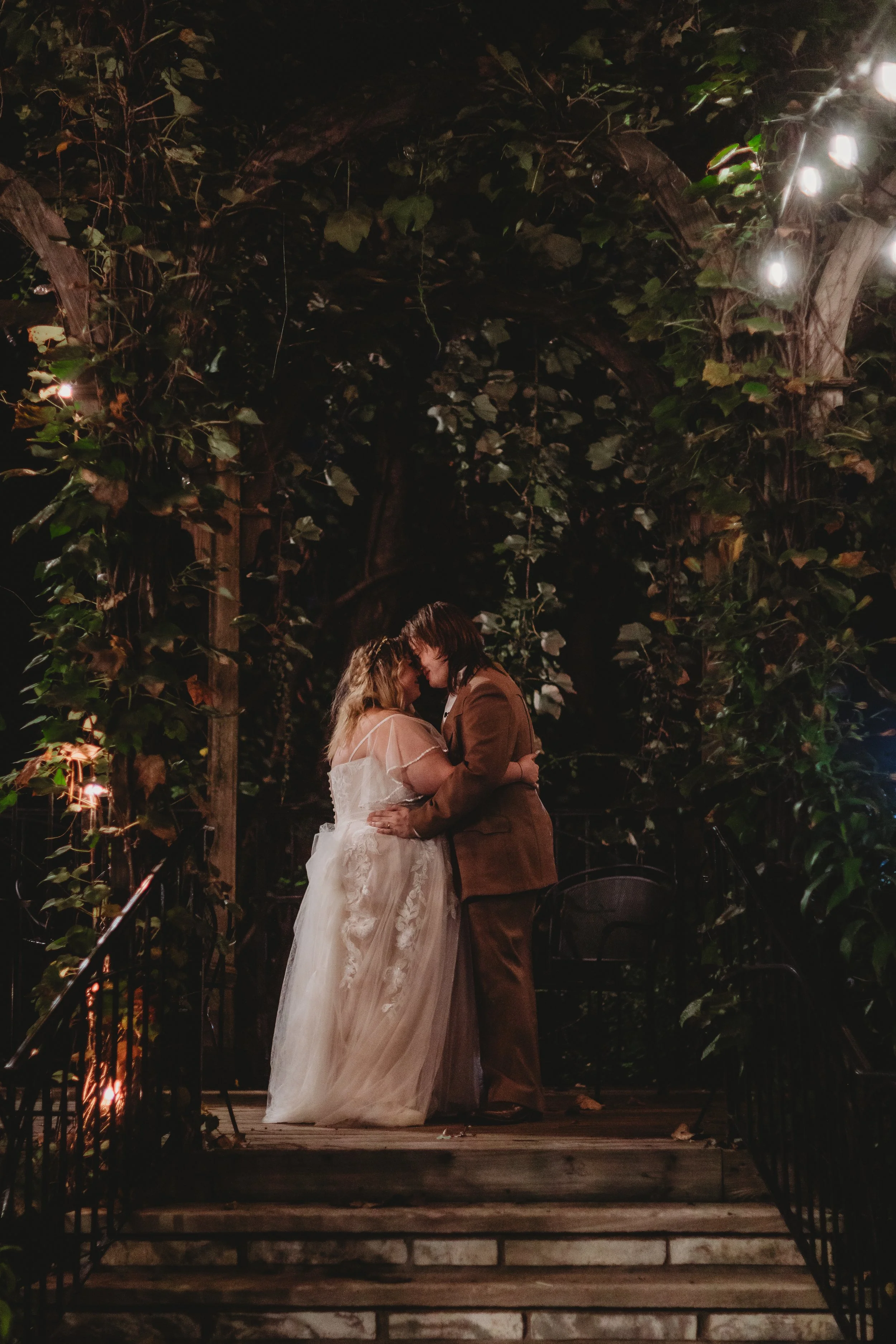 A couple kissing during their wedding ceremony at night, surrounded by greenery and string lights.