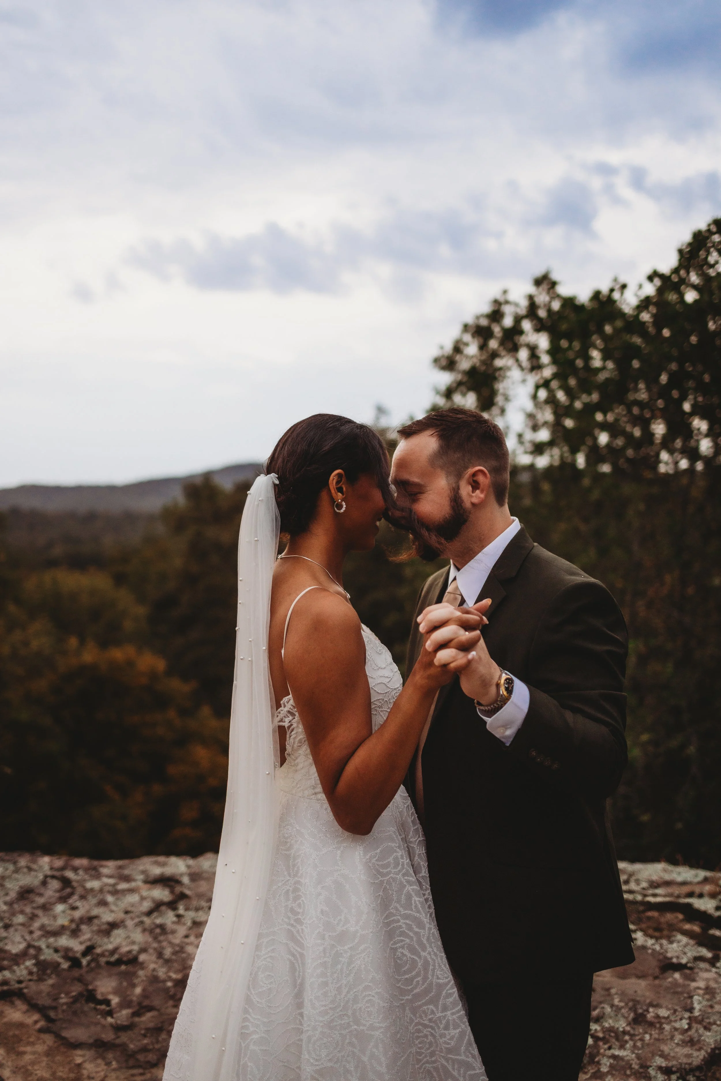 A bride and groom share a dance outdoors during their wedding, holding hands and pressing their foreheads together with a landscape of trees and mountains in the background.