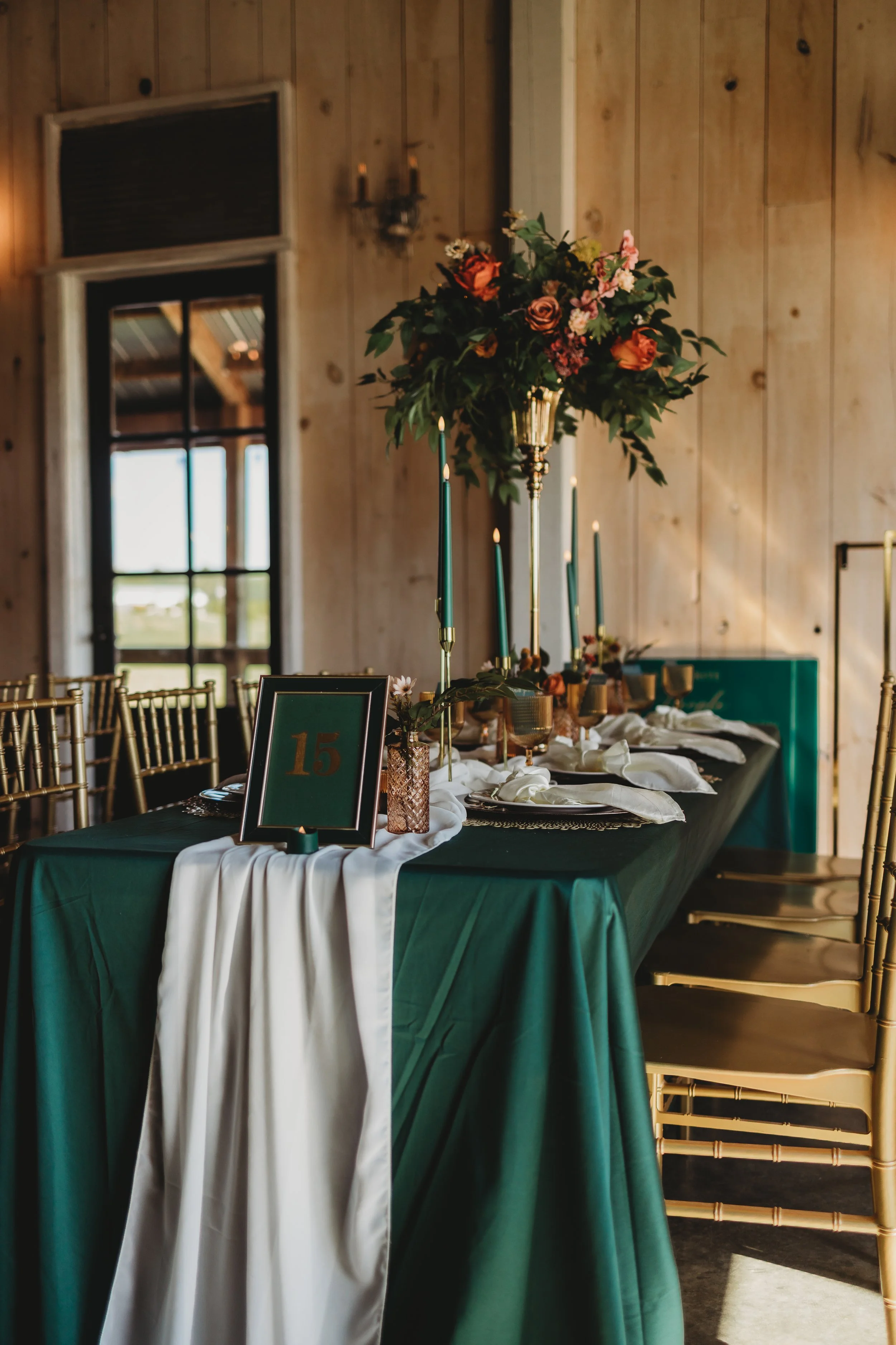 Wedding table decorated with green and white tablecloth, tall floral centerpiece with pink and orange flowers, and gold chairs in a rustic wooden room.