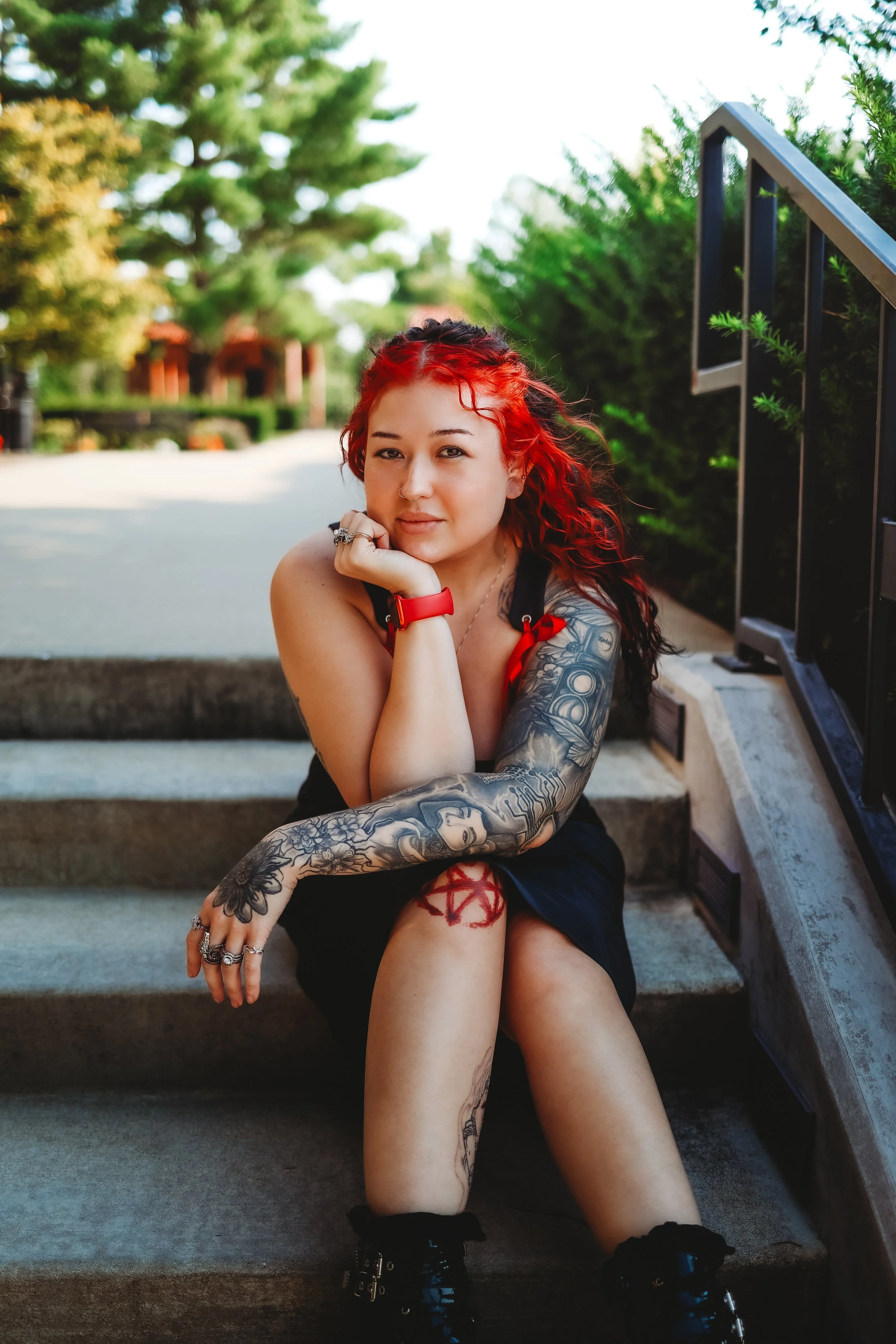 A young woman with red hair, tattoos on her arm and leg, sitting on outdoor stairs, resting her chin on her hand, with a blurred outdoor background of trees and a blue sky.