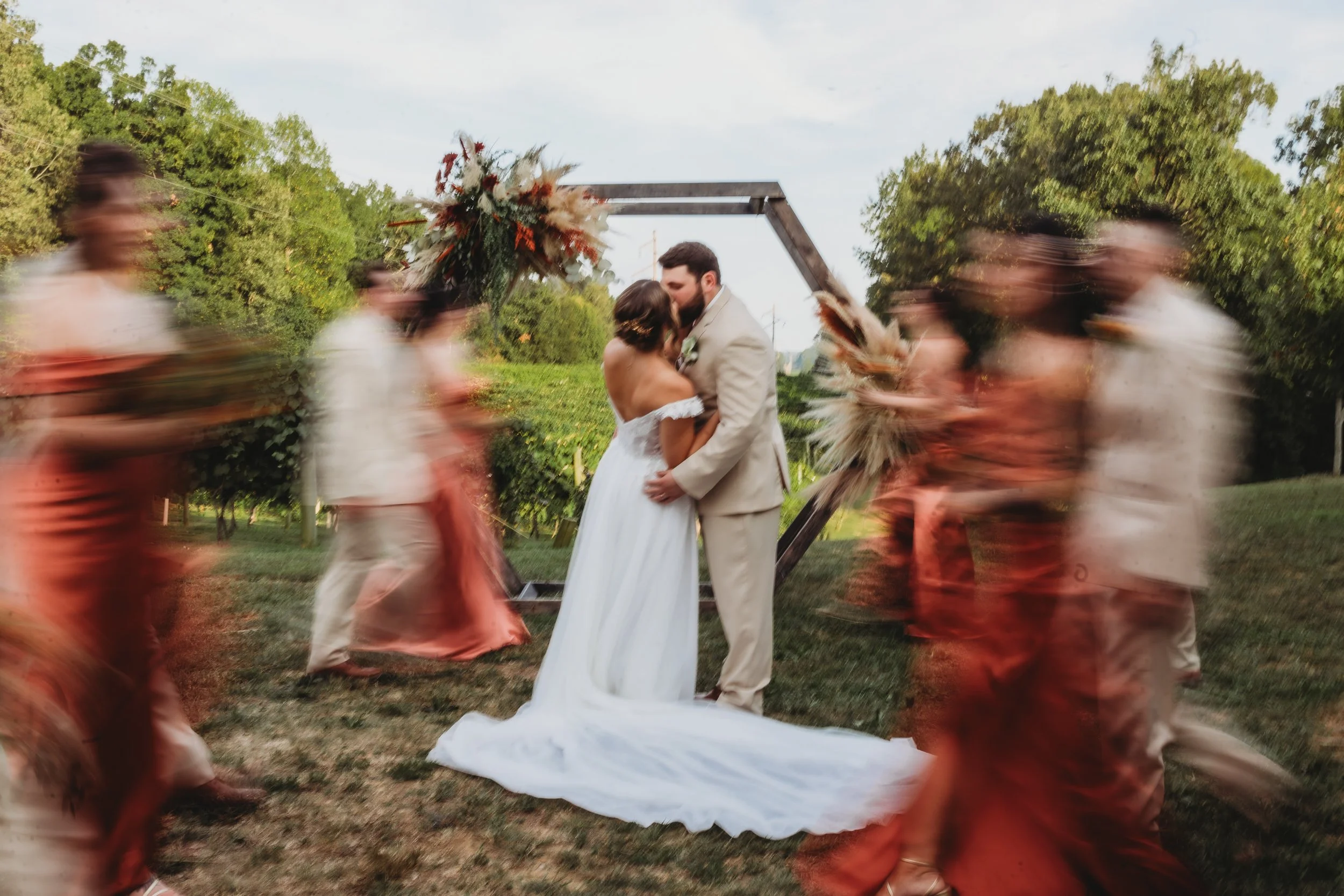 A bride and groom sharing a kiss during their outdoor wedding ceremony surrounded by dancers in pink skirts, with a floral arch and green trees in the background.