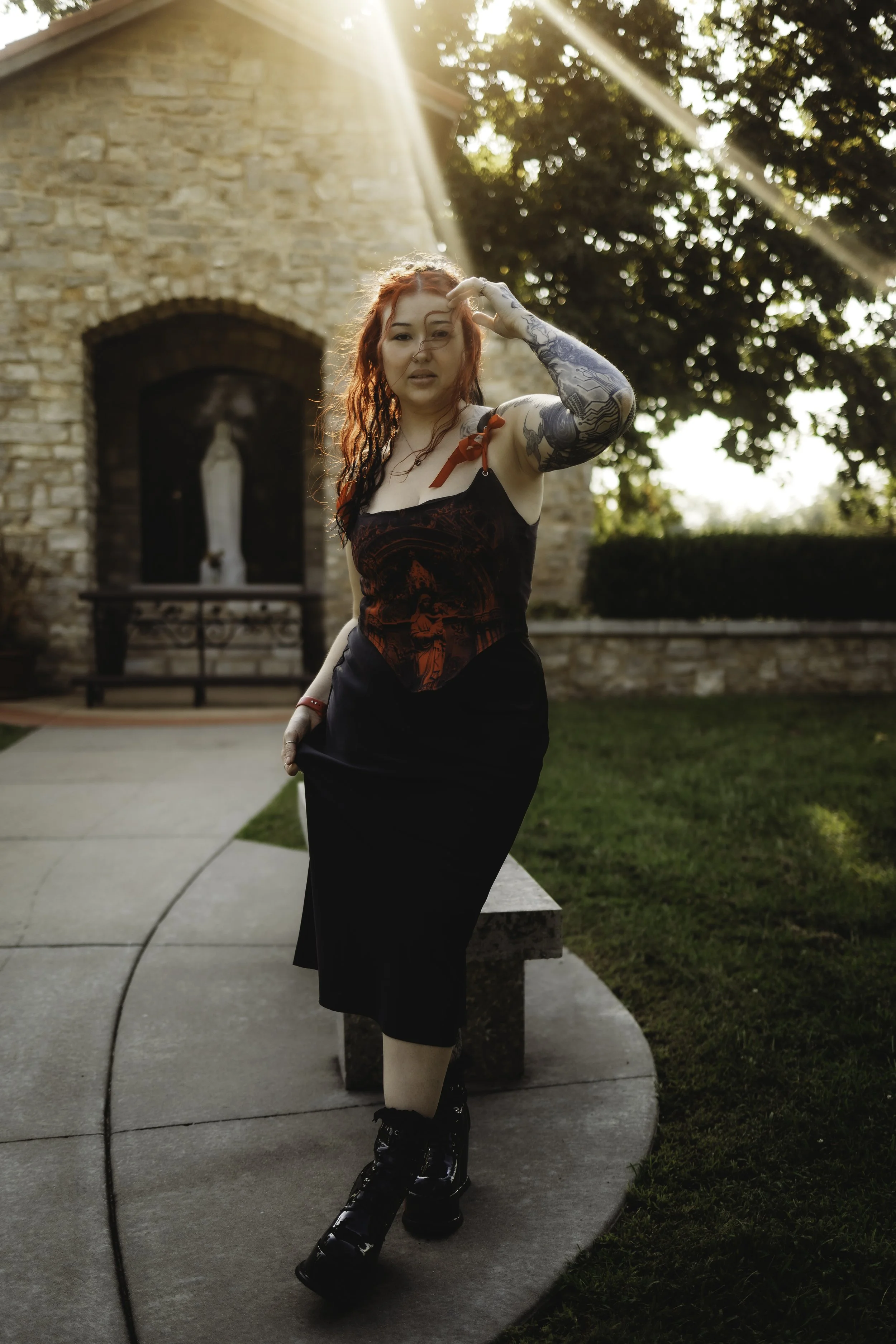 A woman with red hair and tattoos on her arm poses outdoors near a brick building with a religious statue in the background, illuminated by sunlight.