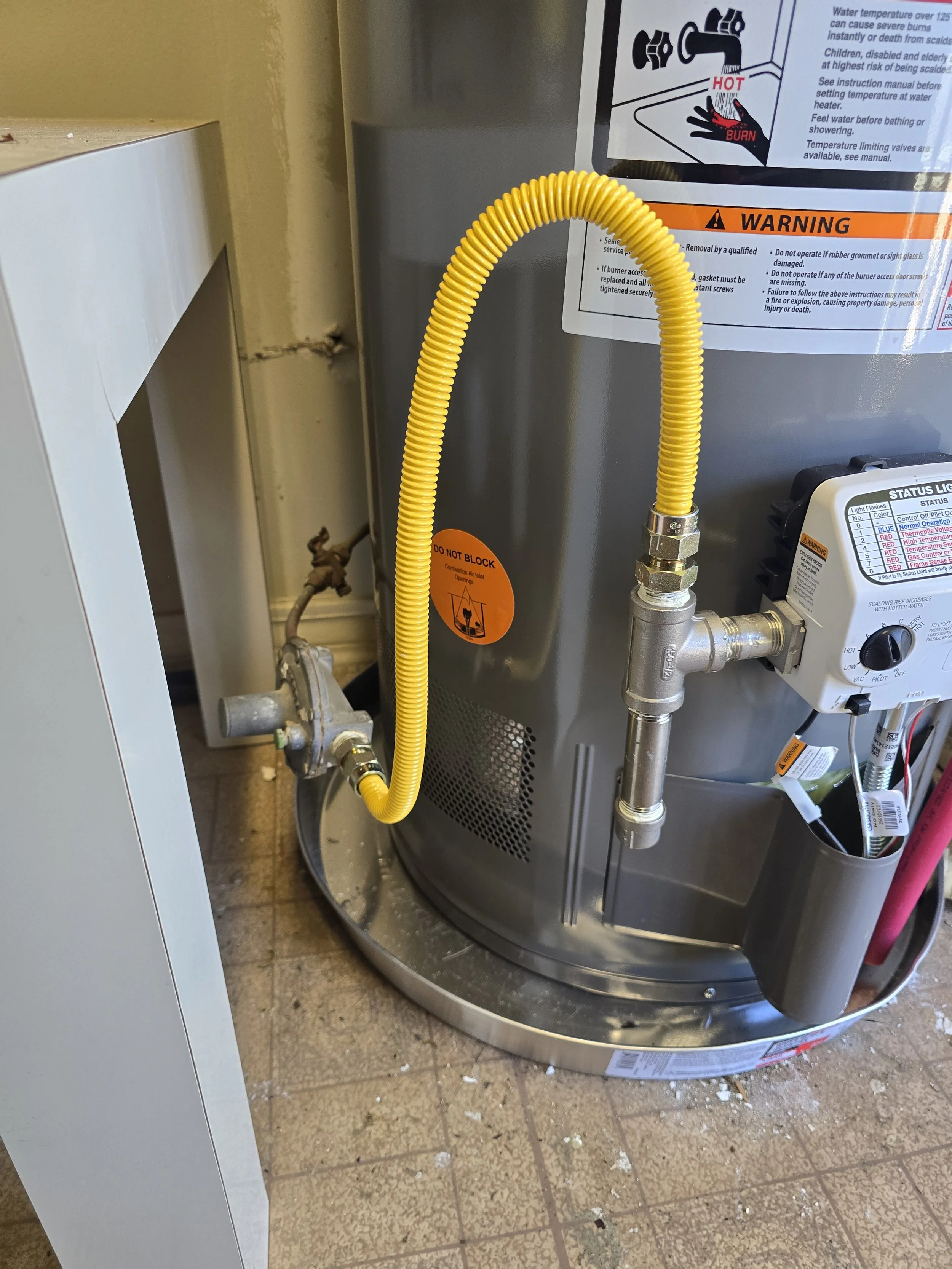 Close-up of a water heater with a yellow flexible gas line, control panel, and warning labels, standing on a tiled floor with debris.
