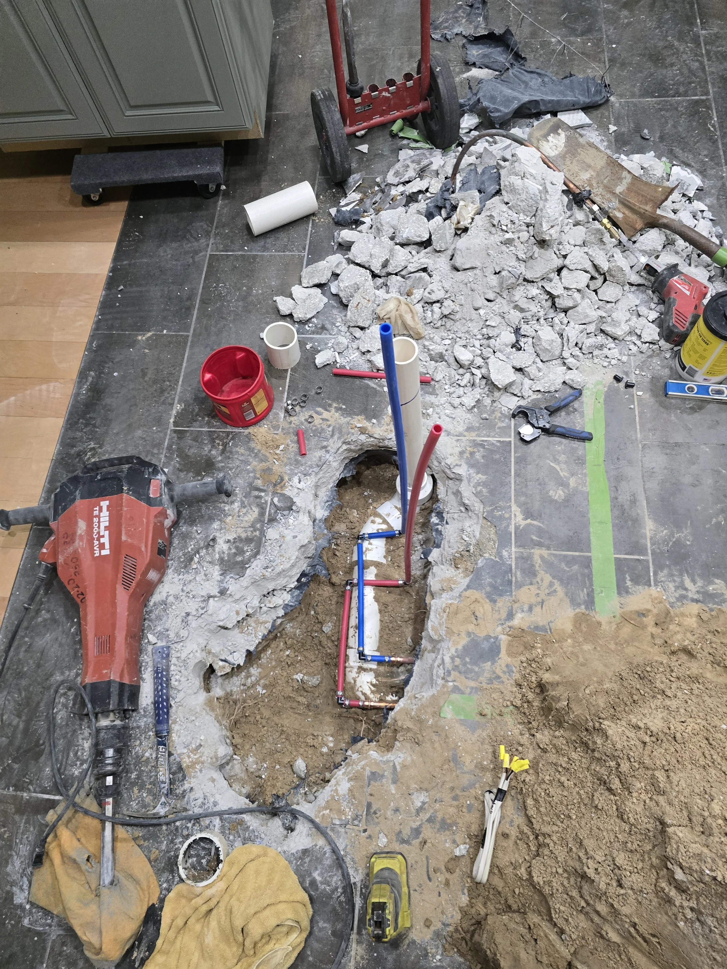 Indoor construction site with broken tile floor, exposed pipes, tools, and debris for plumbing or electrical work.