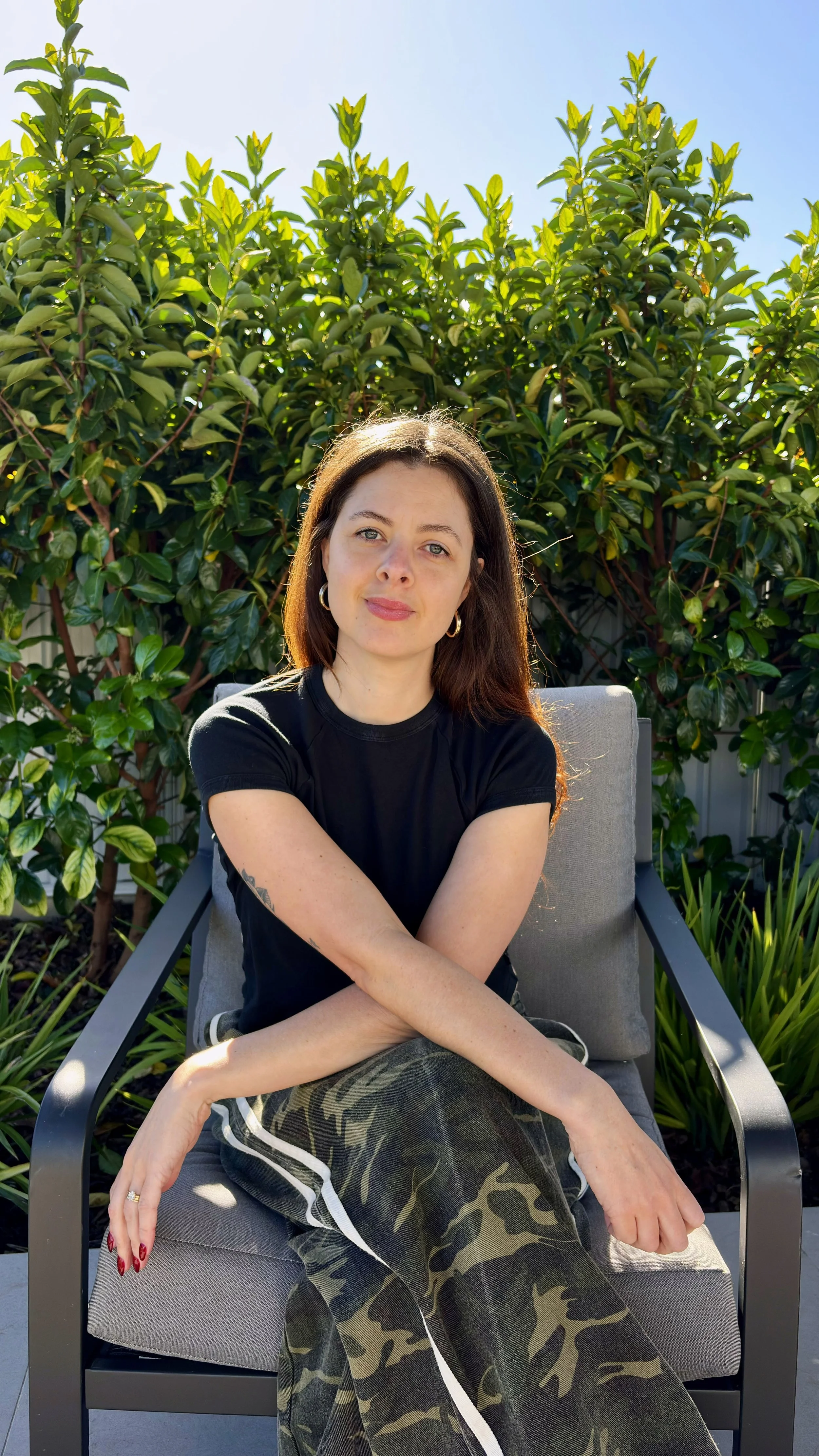 Bon owner of Homeground Marketing with long brown hair, wearing a black t-shirt and camo pants, sitting on a gray outdoor chair with greenery and a clear blue sky in the background.