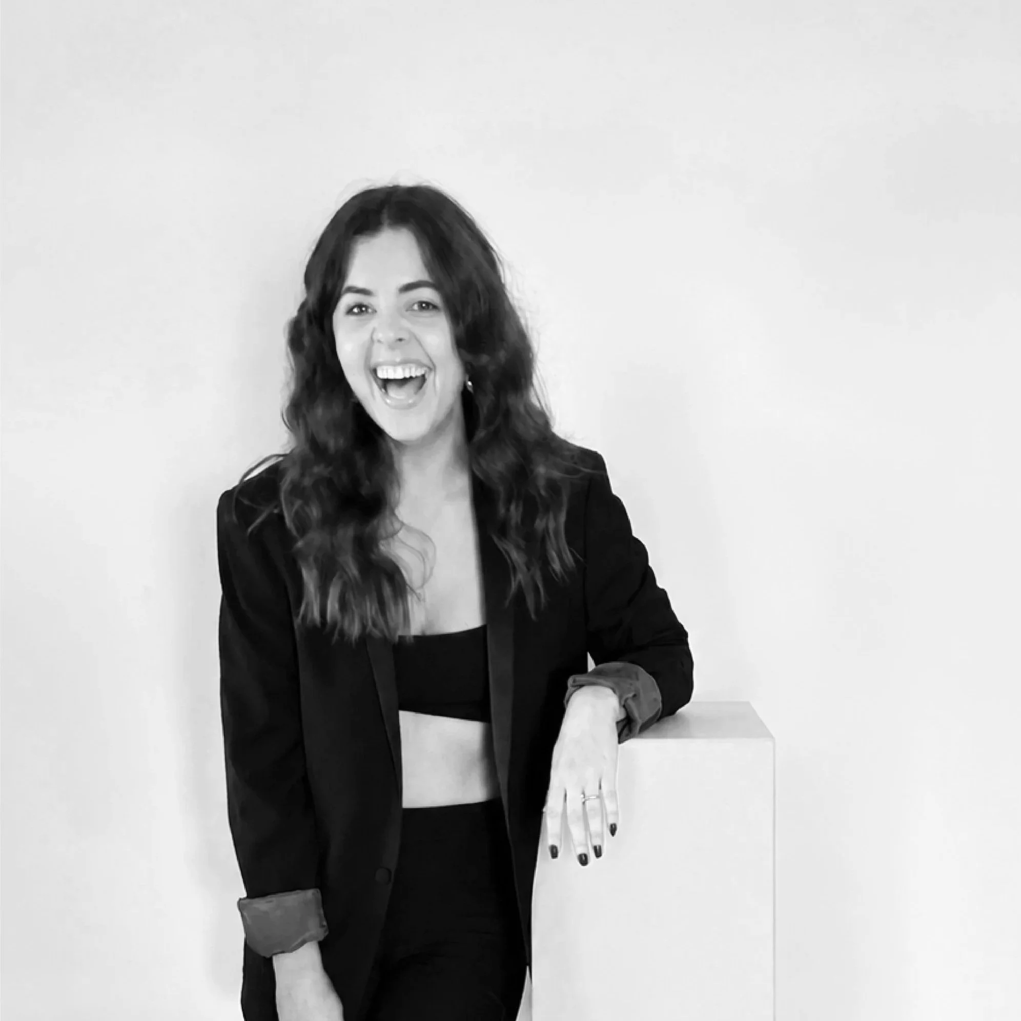 A woman with curly hair and a black blazer, smiling and sitting at a table against a plain wall.