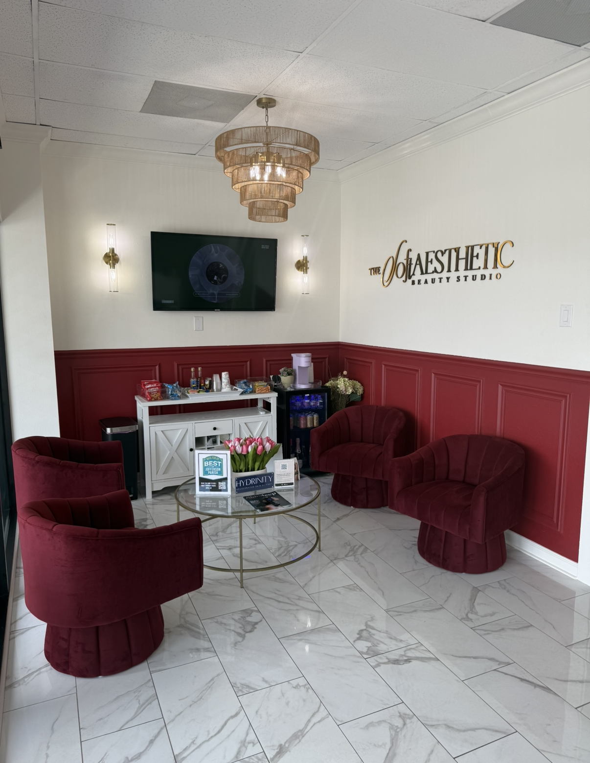 A cozy salon waiting area with four red velvet chairs, a glass coffee table with pink tulips, a white cabinet with snacks and drinks, a small black fridge, and a TV mounted on the wall. The wall features a sign for 'The Aesthetic Beauty Studio,' with gold accents and decorative molding. There is a chandelier hanging from the ceiling and modern wall sconces.