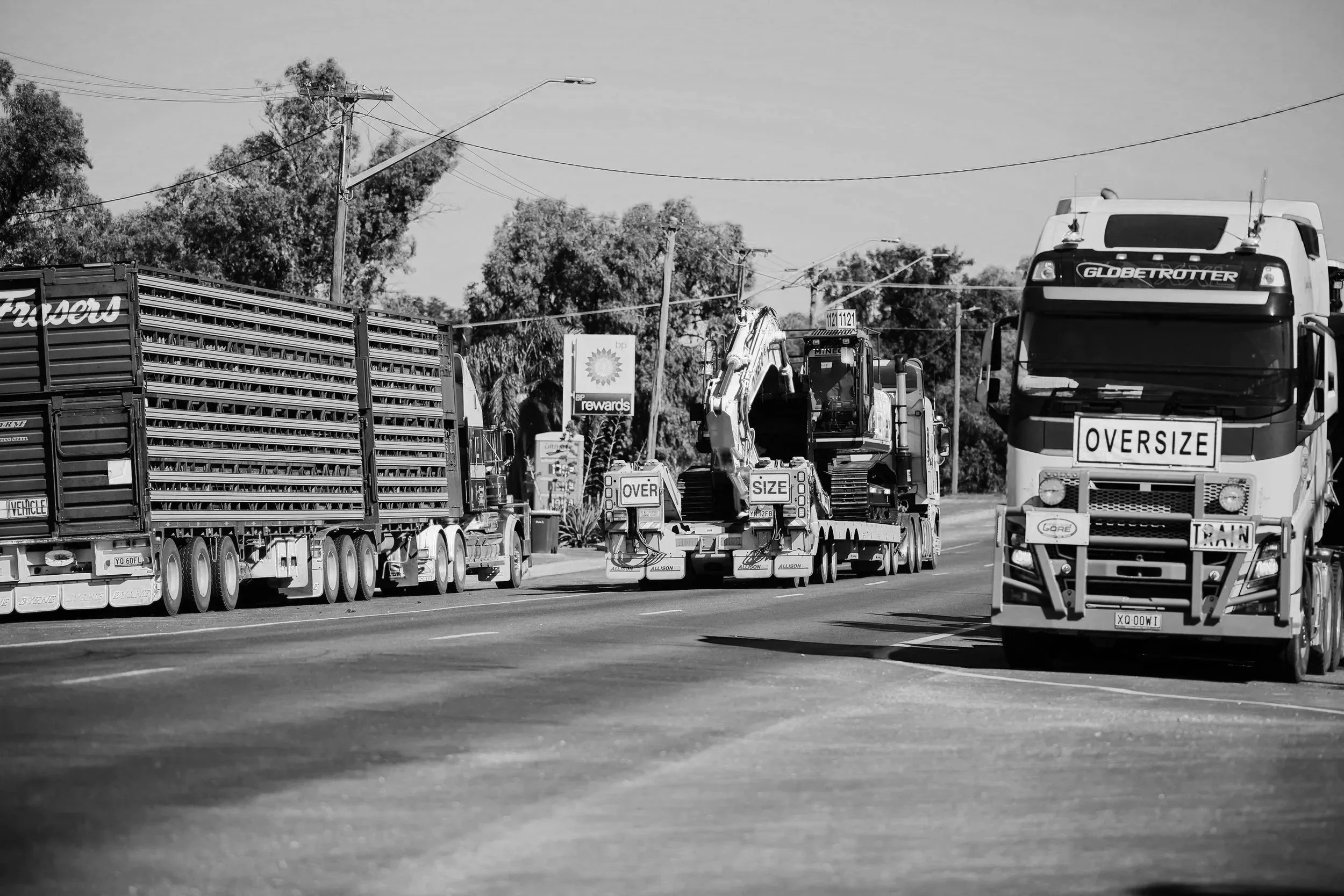 Black and white photo of large trucks and construction vehicles on a highway, with trees and power lines in the background.