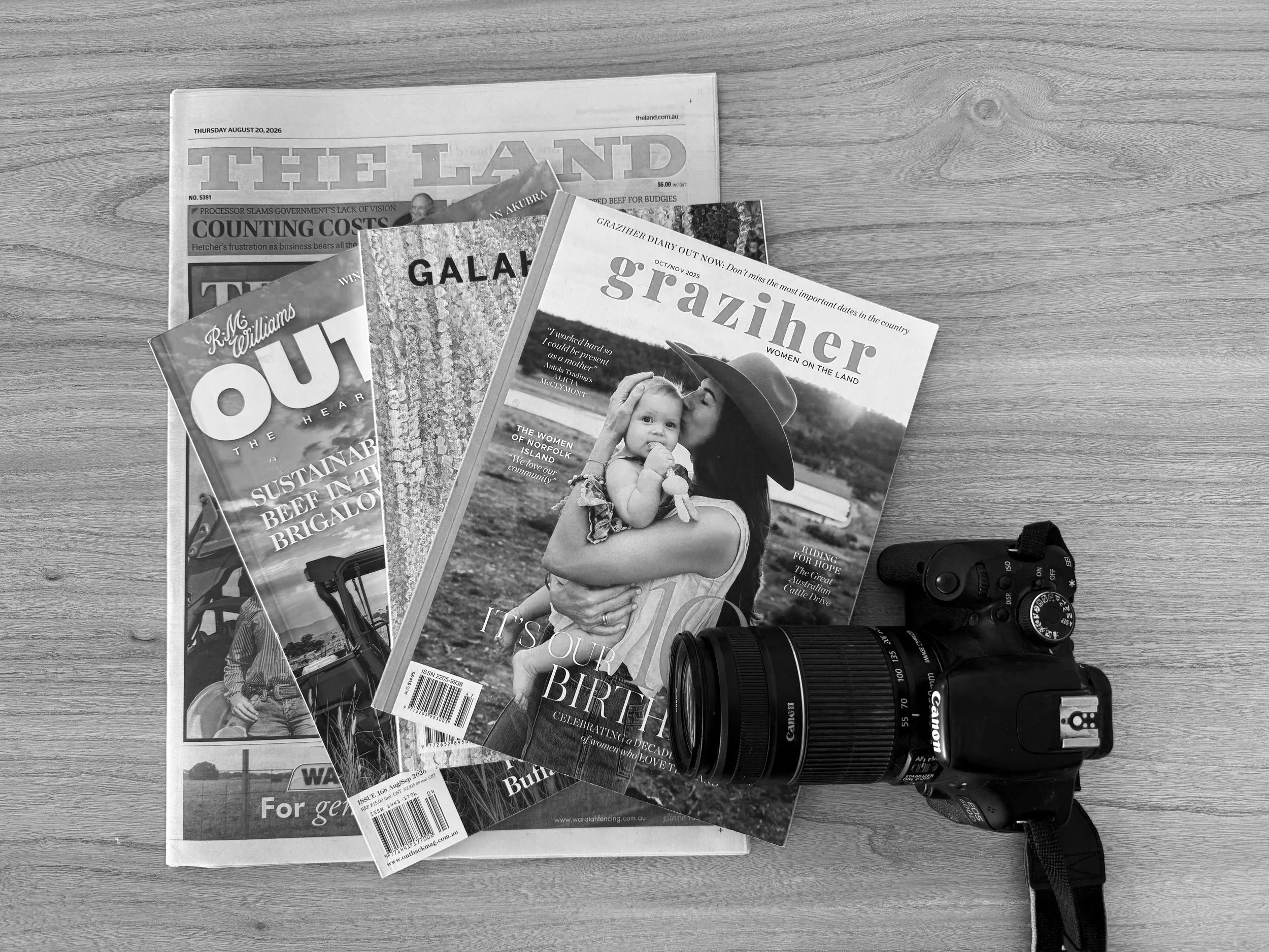 A flat lay of newspapers and magazines on a wooden surface, alongside a Canon camera and lens.