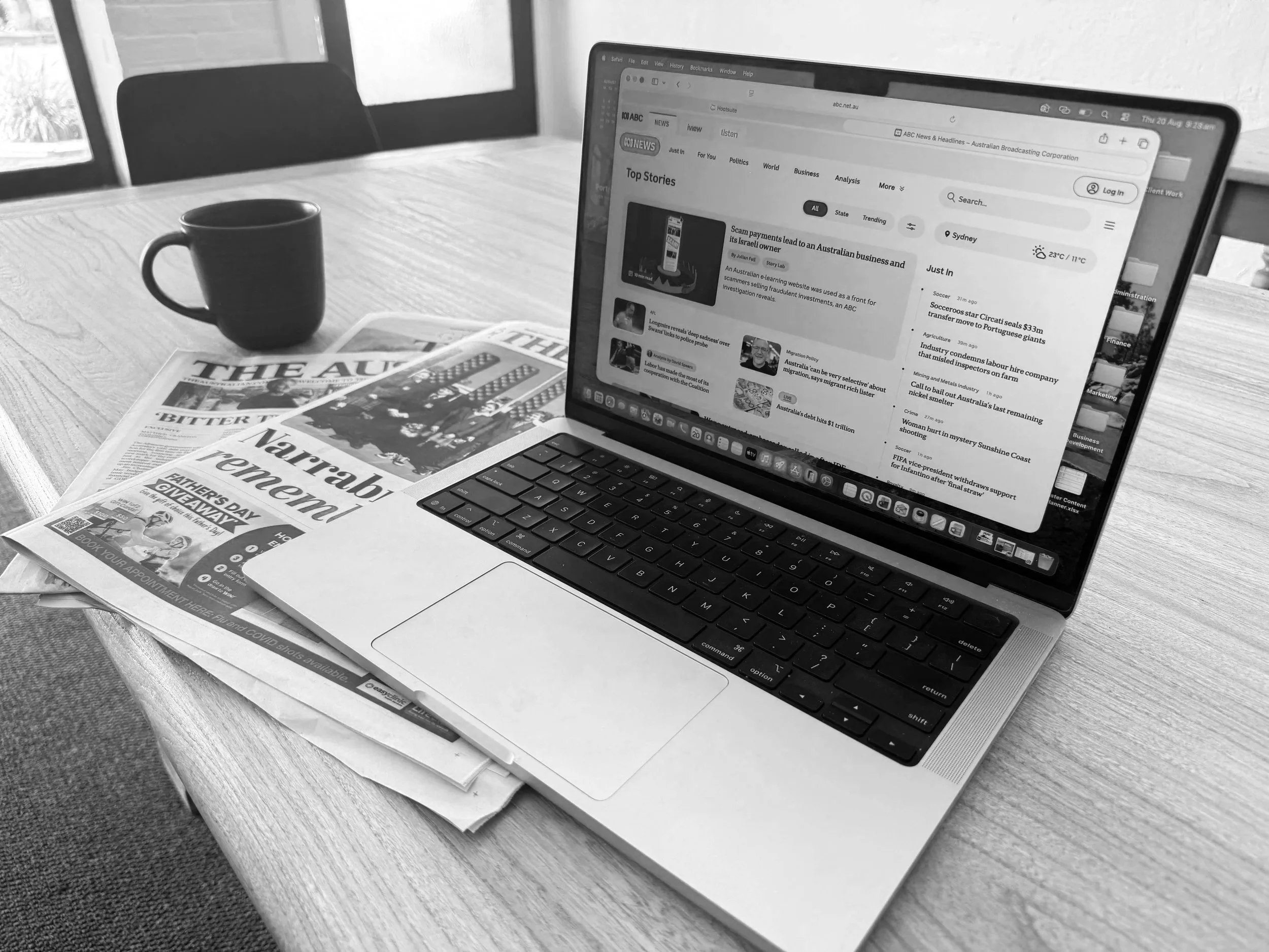 Black and white photo of a laptop on a wooden table with newspapers and a coffee mug nearby, viewed near a window.