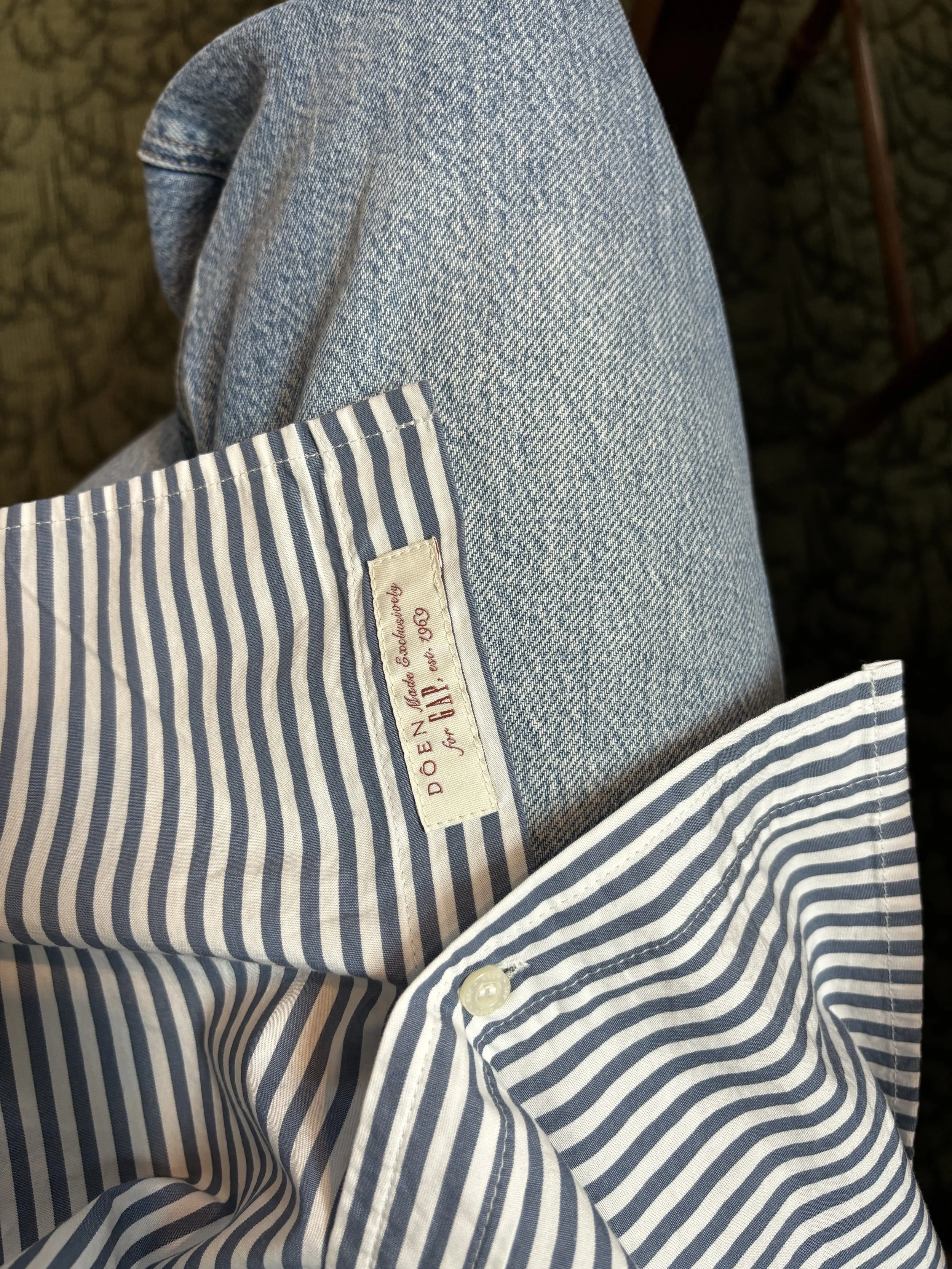 Close-up of a blue and white striped shirt with a white button and a beige label that reads 'DOR..'. A person is wearing light blue jeans. The image is taken from above, focusing on the person's lap and the shirt.