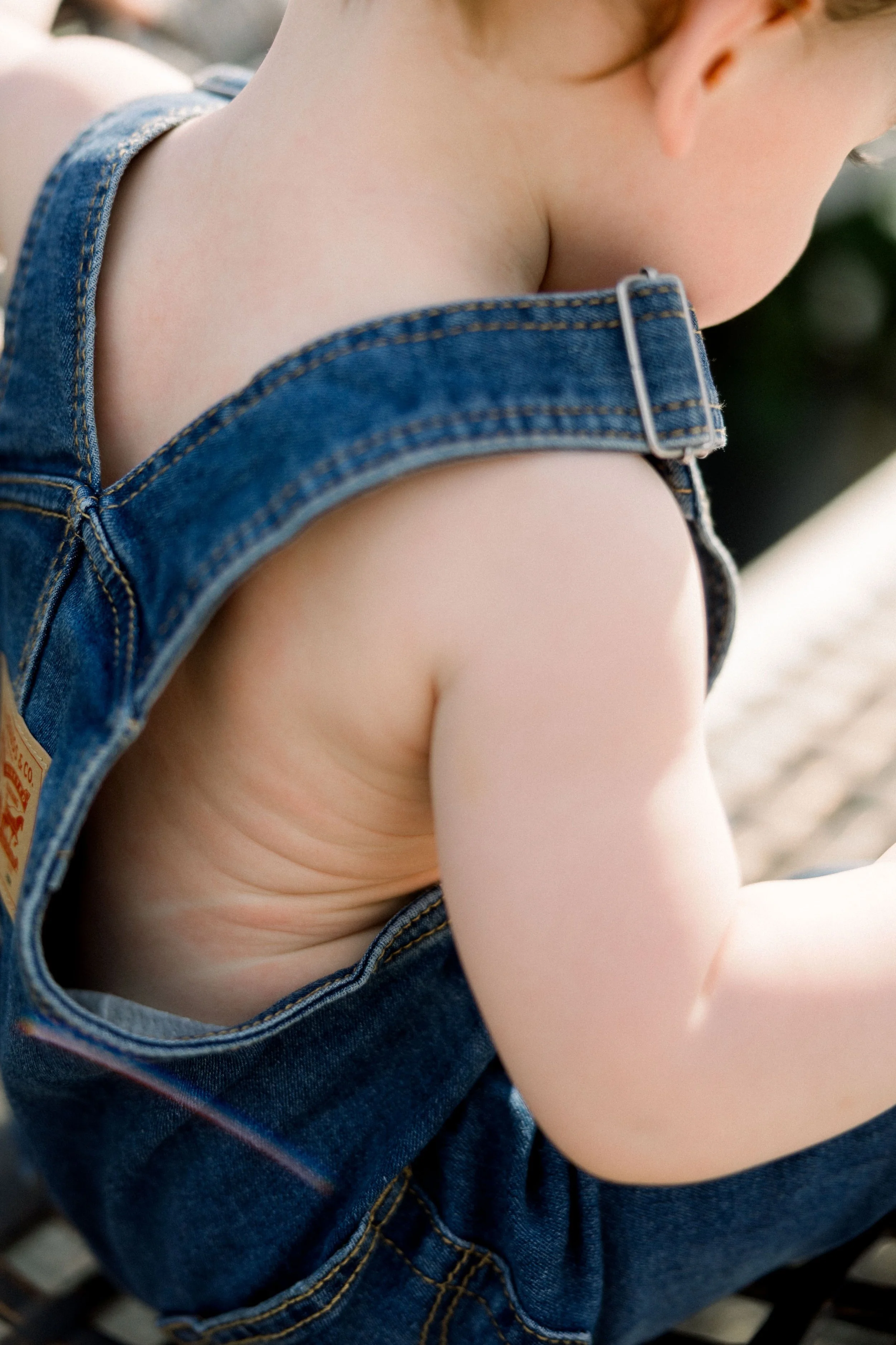 Close-up of a young child wearing denim overalls, showing their shoulder, arm, and part of their face and hair.