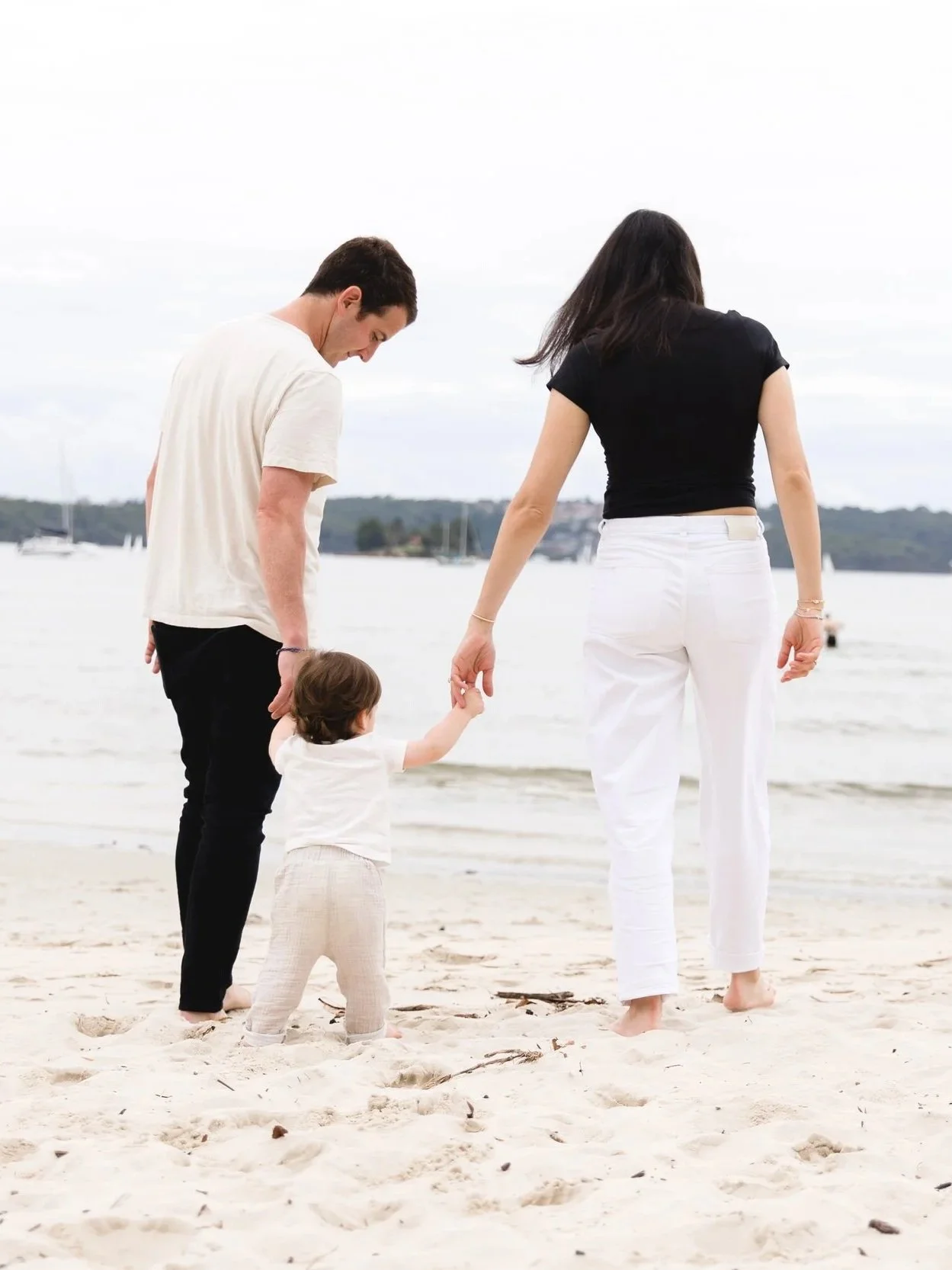 A family walking on the beach with a man, woman, and young girl holding hands and facing away from the camera, with sailboats in the background.
