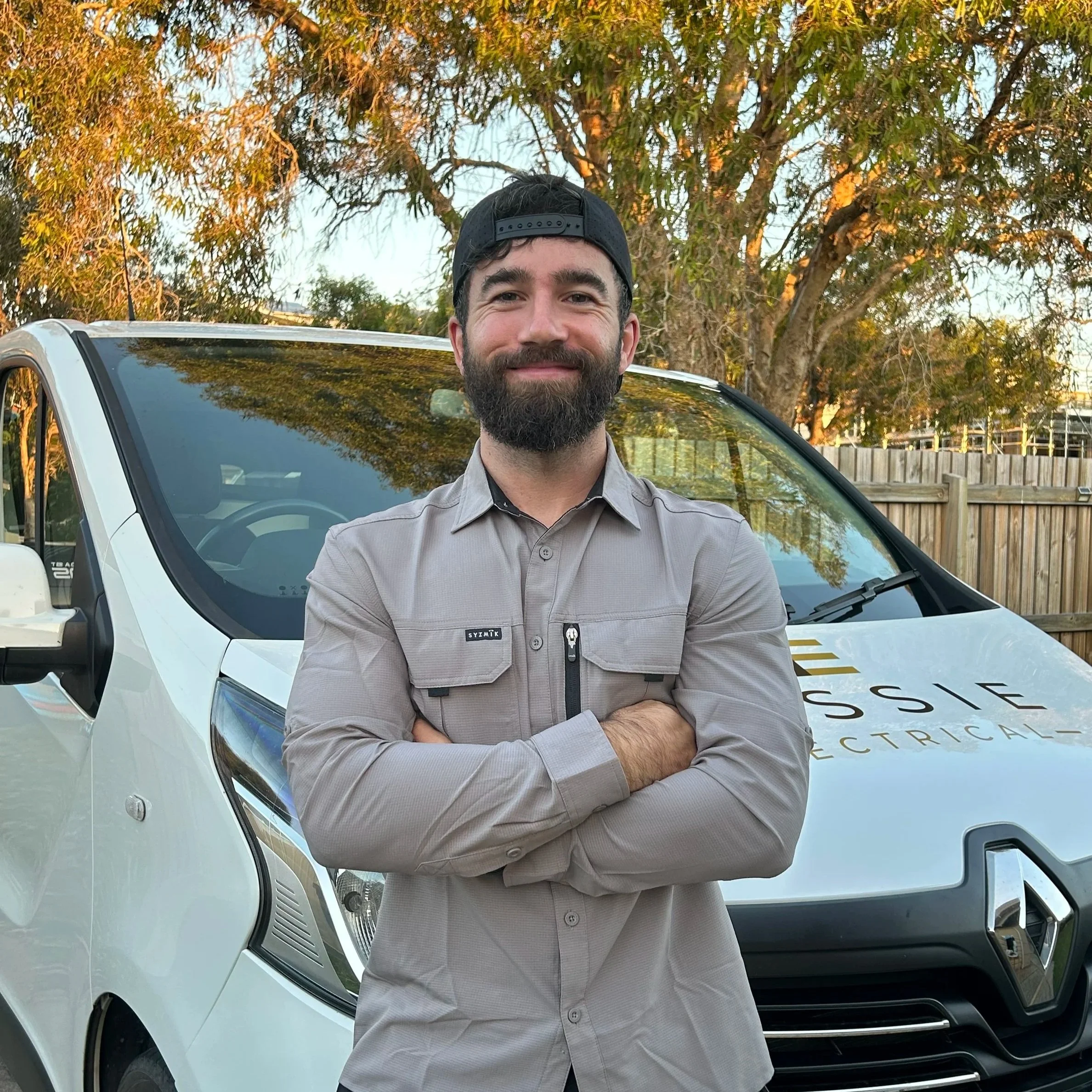 A man with a beard wearing a gray button-up shirt and a backwards black cap, standing outdoors in front of a white vehicle with a tree and fence in the background.