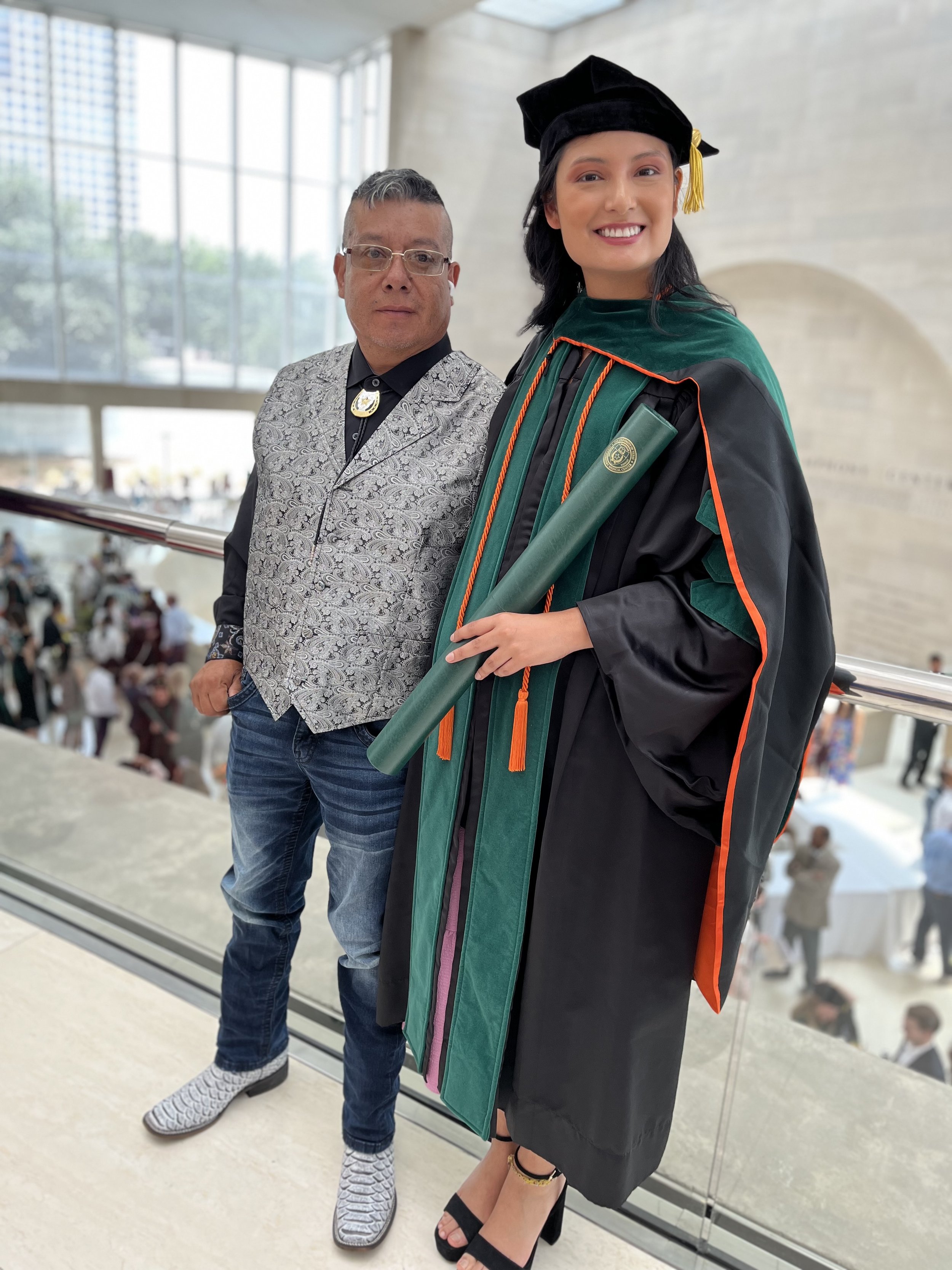 A woman in graduation regalia holding a diploma standing beside a man in casual attire, both in an indoor setting with large windows and a crowd in the background.