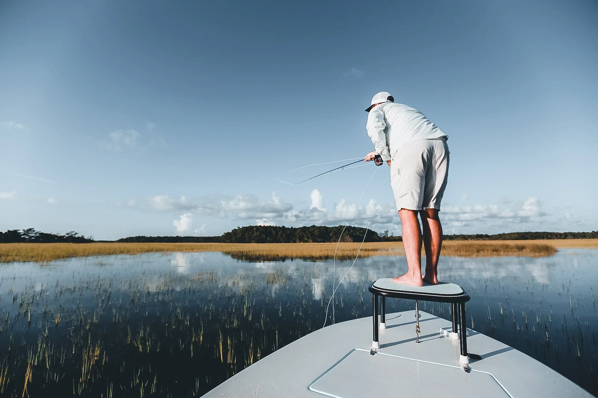 Person standing on a boat fishing in a marshland with a blue sky and scattered clouds.