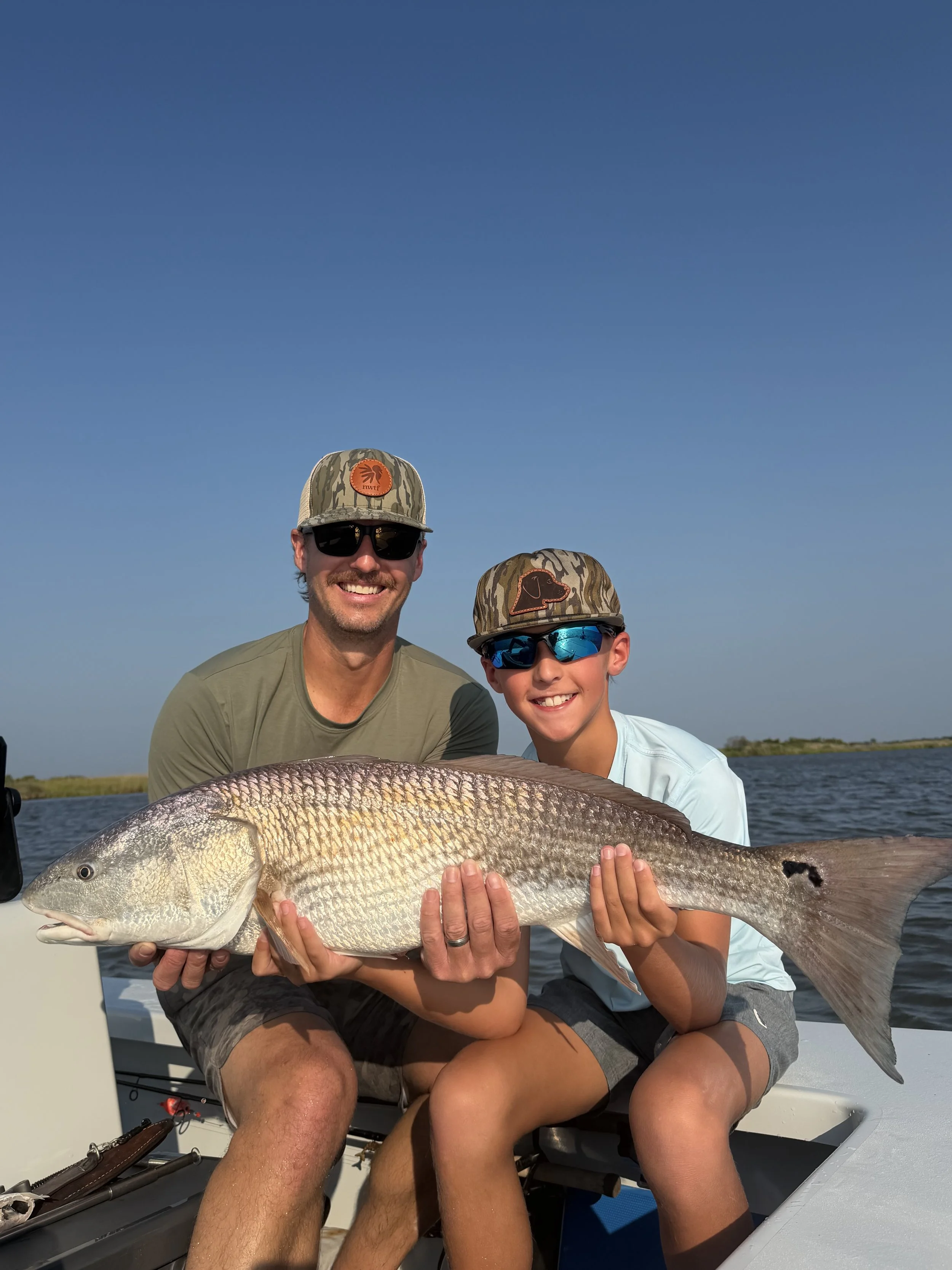 Two males sitting on a boat holding a large fish, wearing camouflage caps and sunglasses, smiling against a backdrop of water and blue sky.