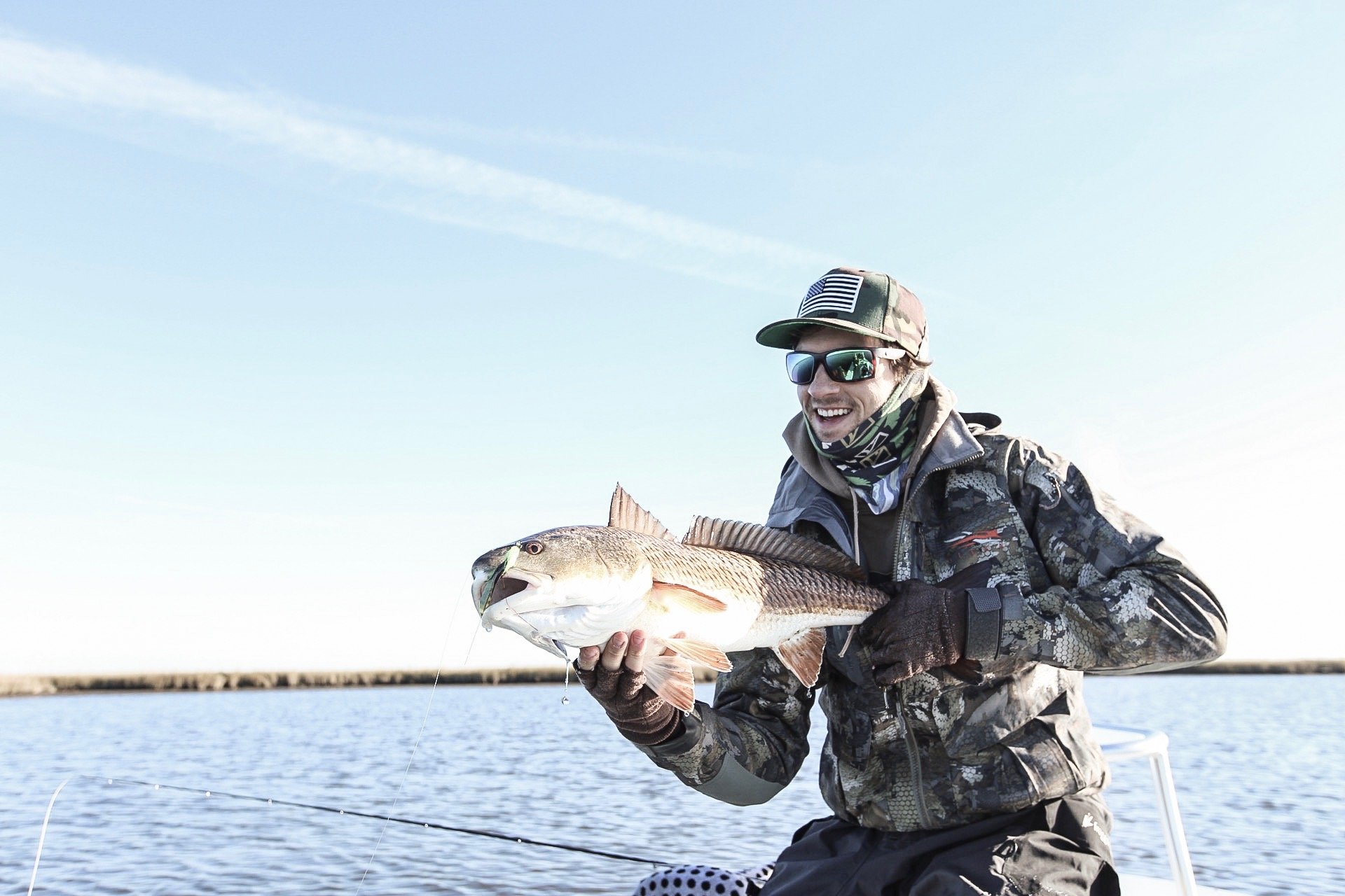 A man in camouflage outdoor clothing, sunglasses, and a baseball cap with an American flag patch, is smiling and holding a large fish on a boat in a body of water under a clear sky.