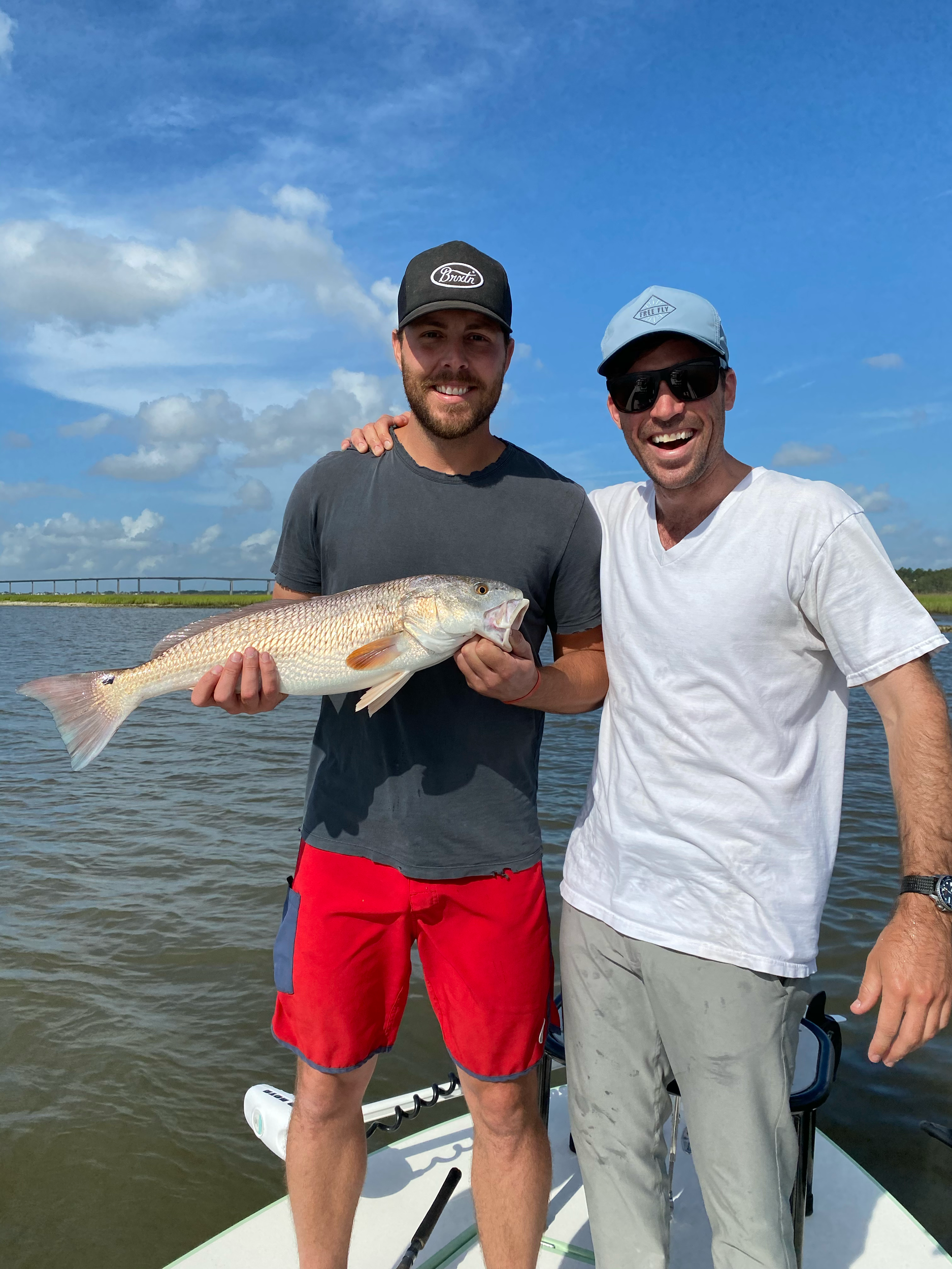 Two men on a boat holding a large fish, smiling, with a blue sky and water in the background.