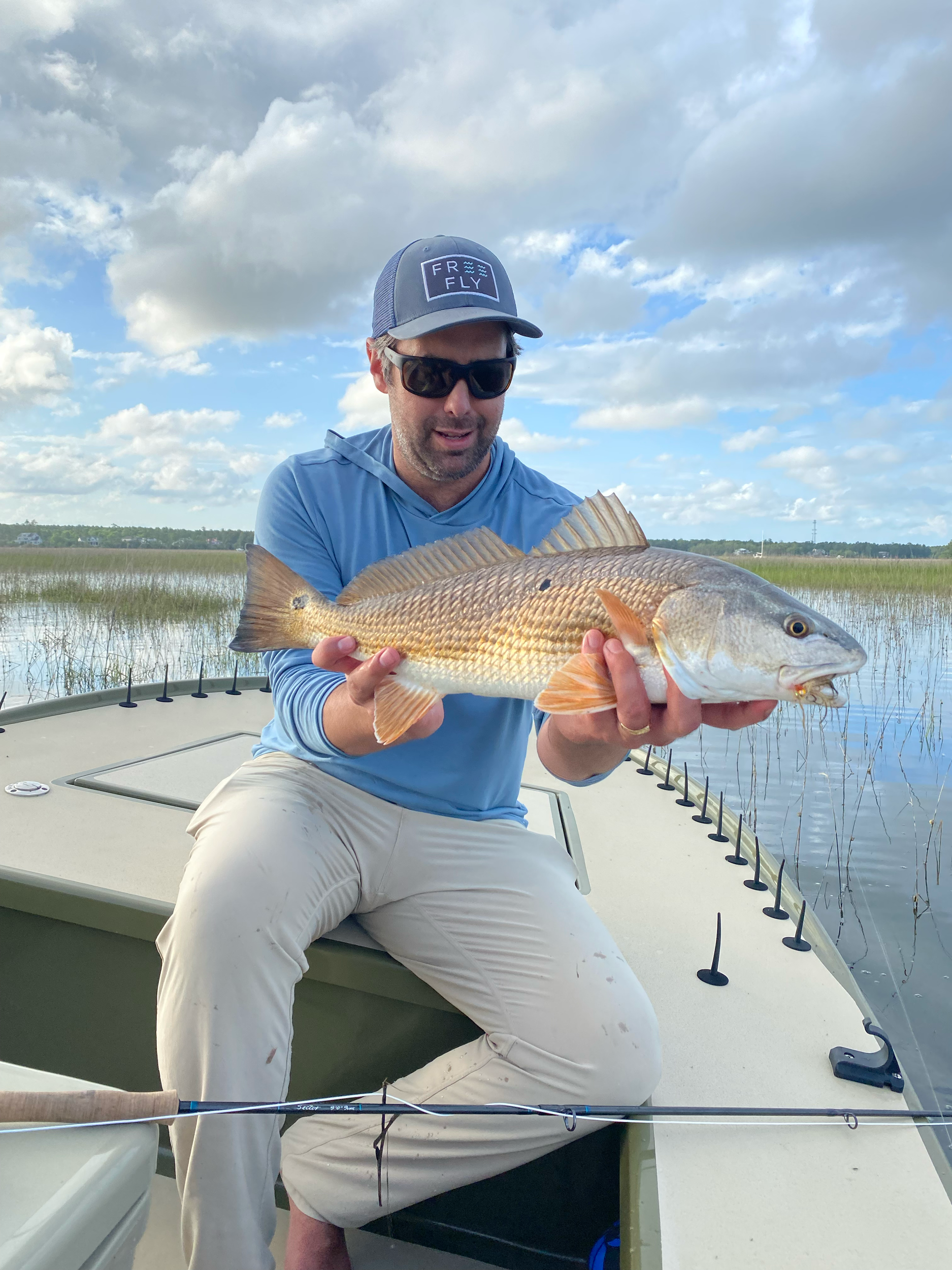 A man holding a large fish on a boat with a wetland and cloudy sky in the background.