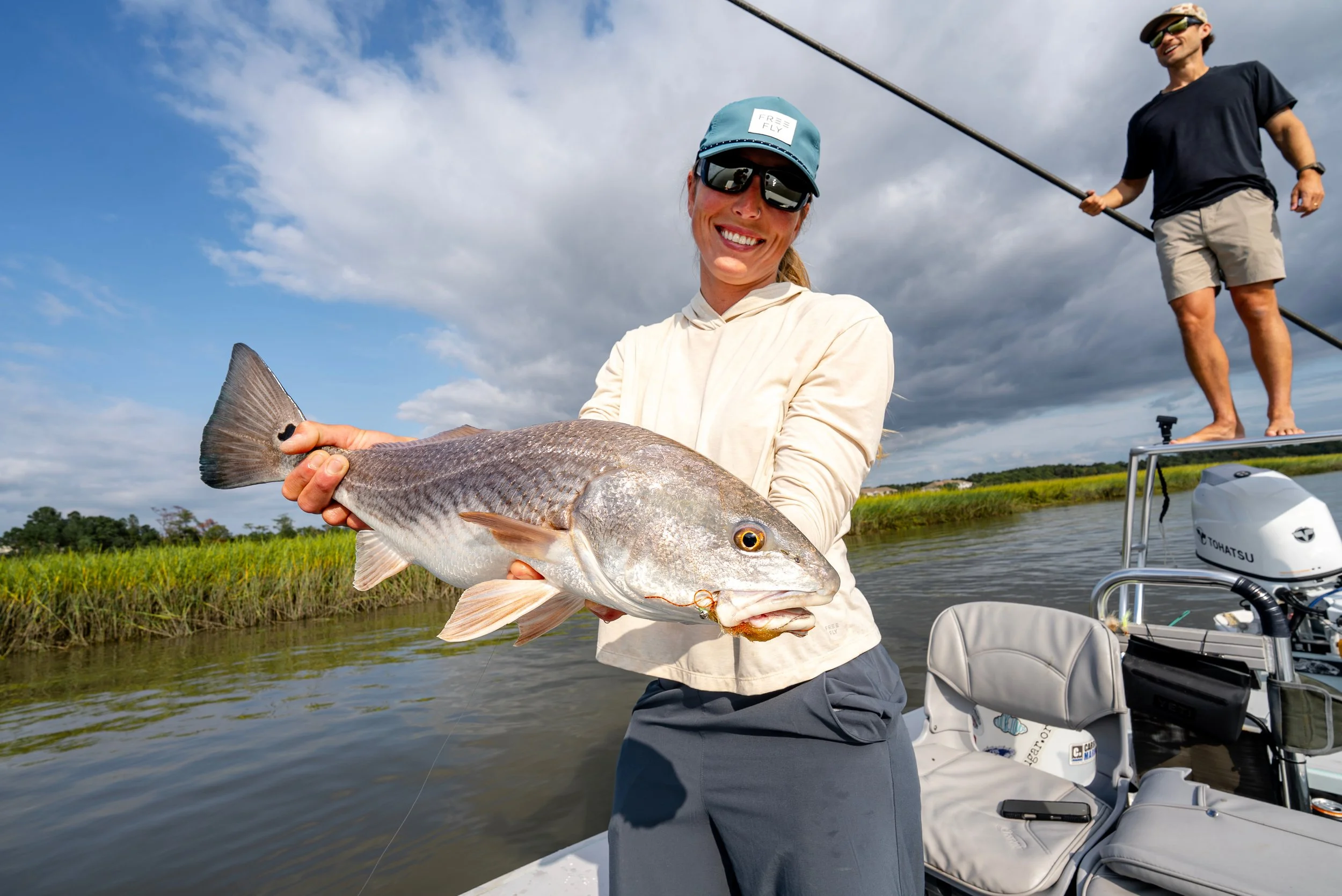 A woman on a boat holding a large fish she caught, with a man in the background on the boat holding a fishing rod and smiling, under a partly cloudy sky near a river or lake.