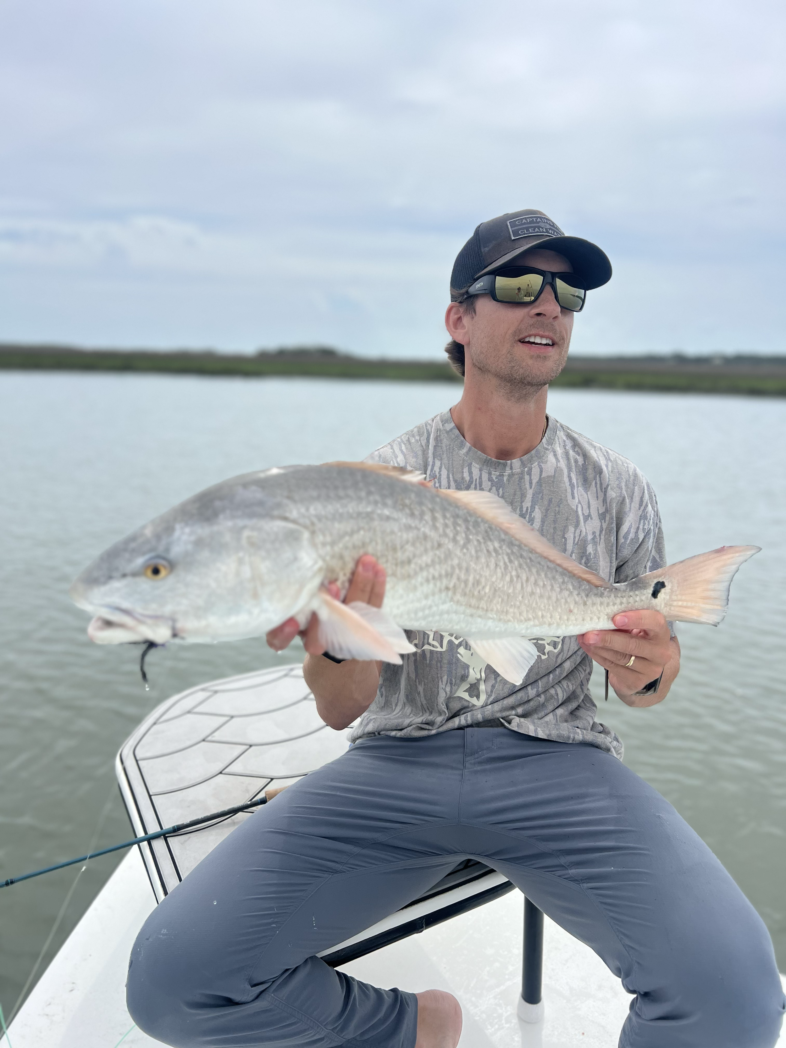 A man in a gray t-shirt, dark sunglasses, gray pants, and a black cap sitting on a boat holding a fish over the water, with a landscape of water and distant land in the background.