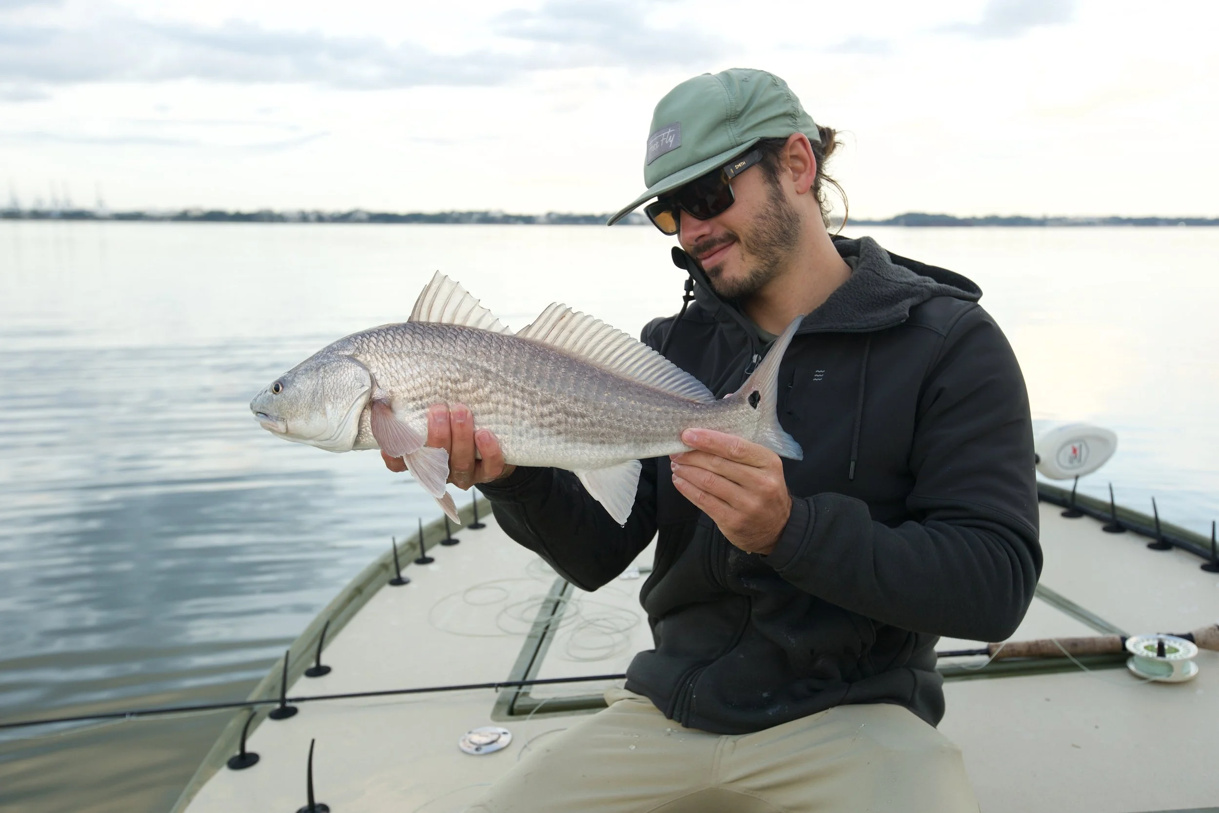 A man holding a large fish on a boat in a body of water, wearing sunglasses, a hat, and a dark jacket.