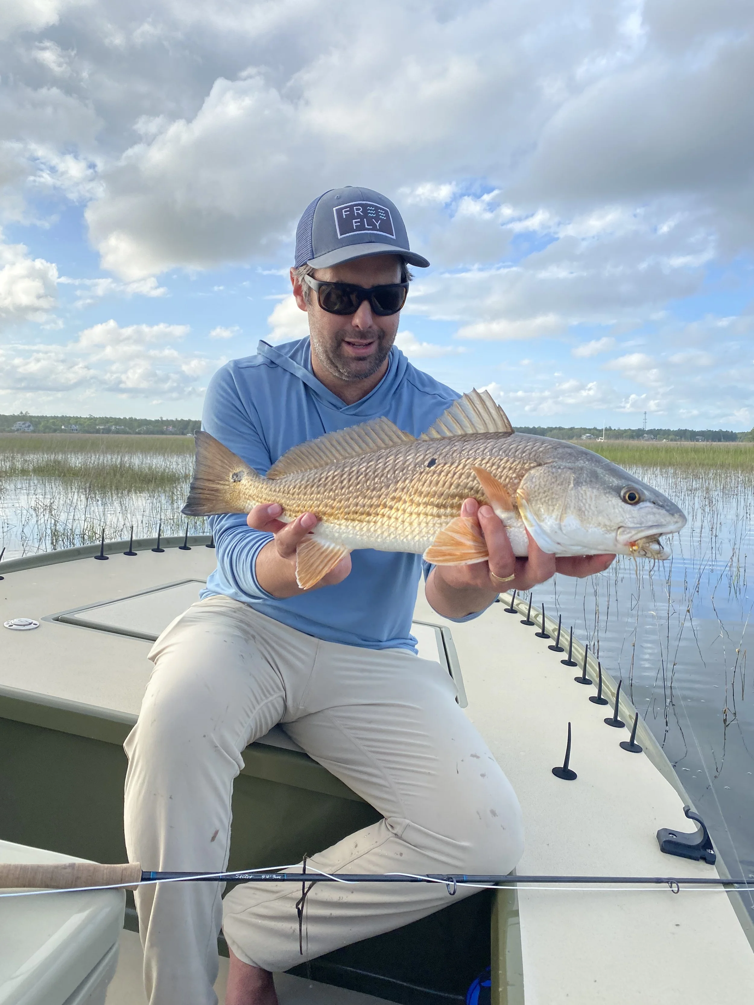 A man wearing sunglasses and a blue cap with 'FRO FLY' on it, seated in a boat, holding a large fish, with a background of water, grass, and a partly cloudy sky.