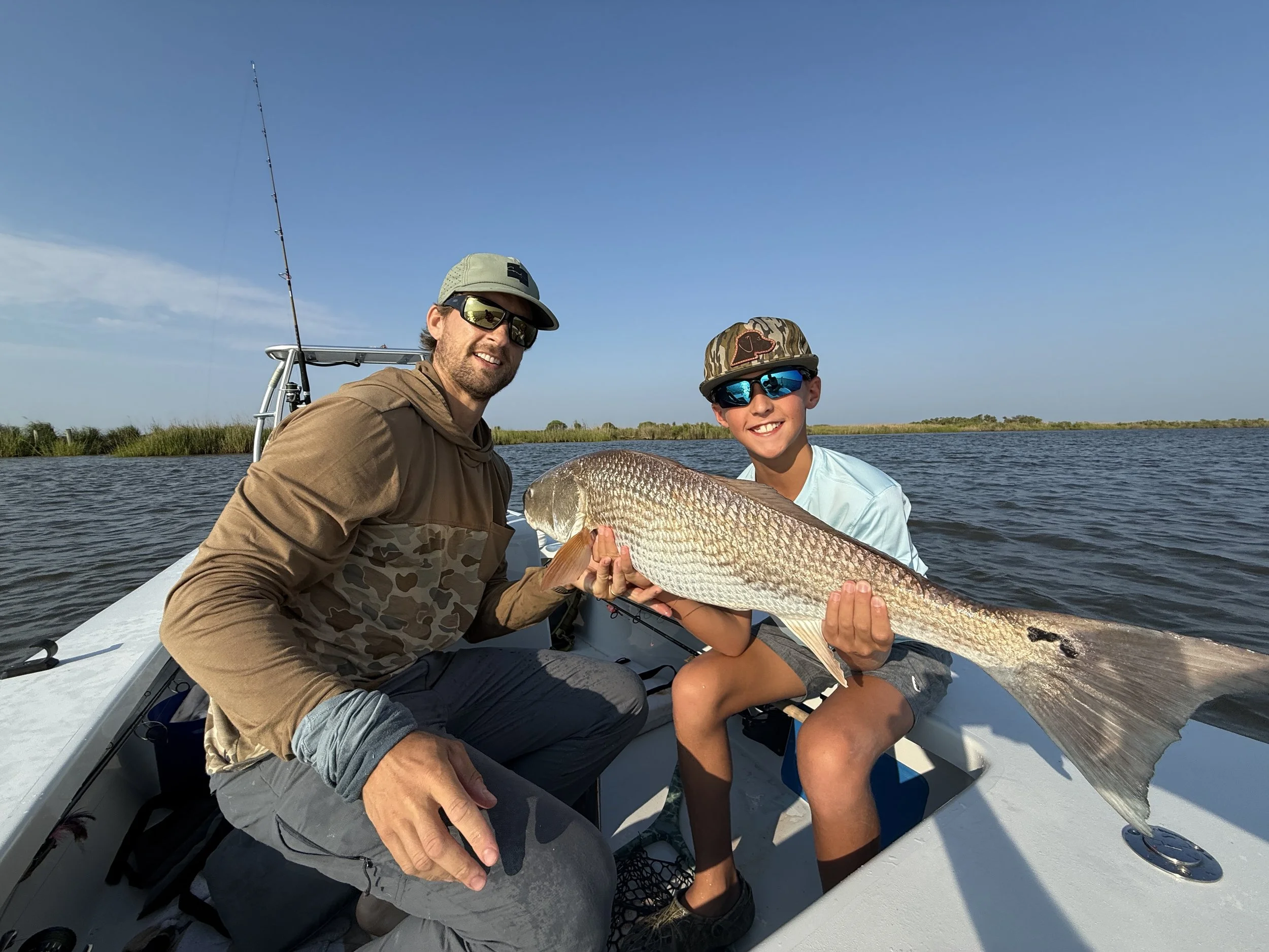 A smiling man and boy holding a large fish while sitting on a boat in a waterway during daytime.