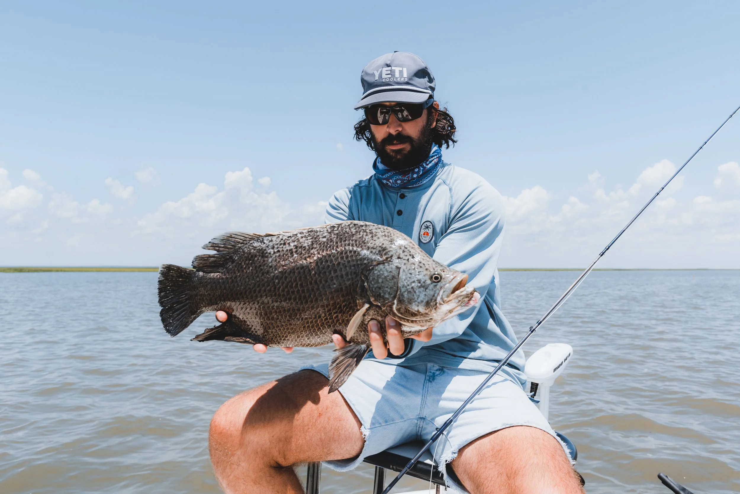 A man wearing sunglasses, a gray cap, and a blue long-sleeve shirt sitting on a boat, holding a large fish over the water.