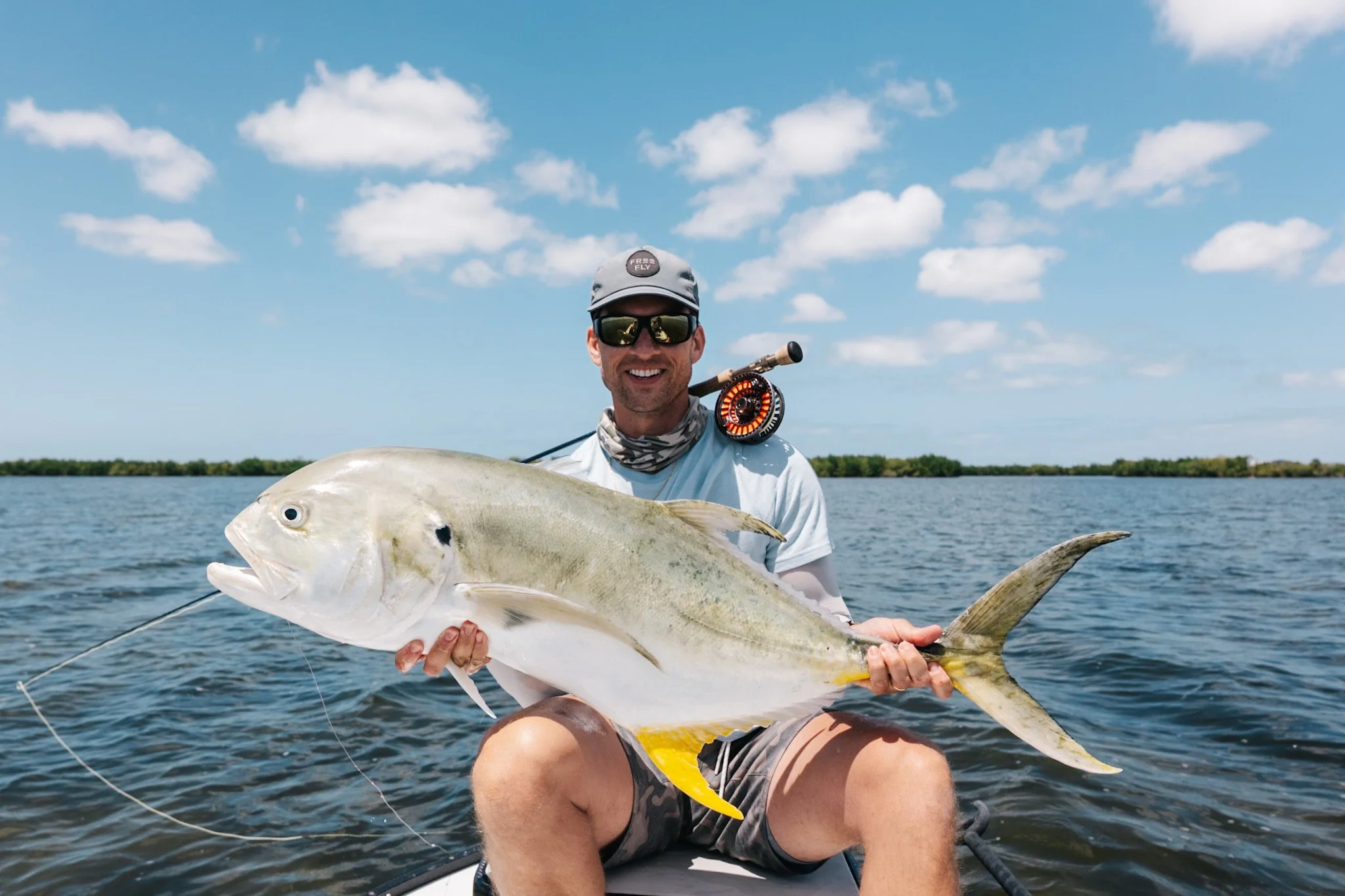 A man smiling and wearing sunglasses, a gray cap with a patch reading 'FREE FLY', and a neck gaiter, holding a large white fish with pale coloring. He is on a boat in a body of water with a distant shoreline visible and a partly cloudy sky overhead.