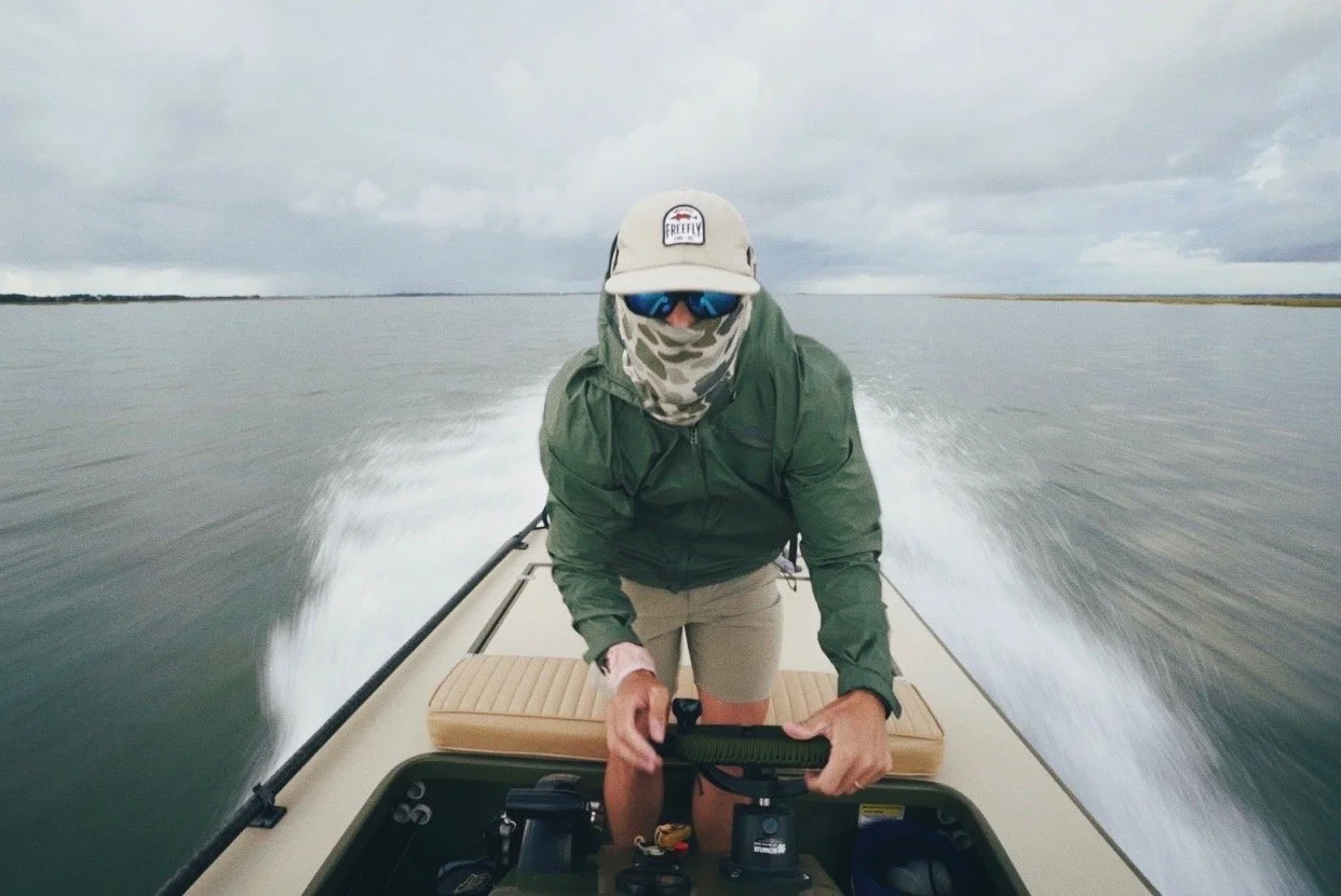 Person wearing sunglasses, a cap, face covering, of green jacket, navigating a small boat on a calm, overcast lake.