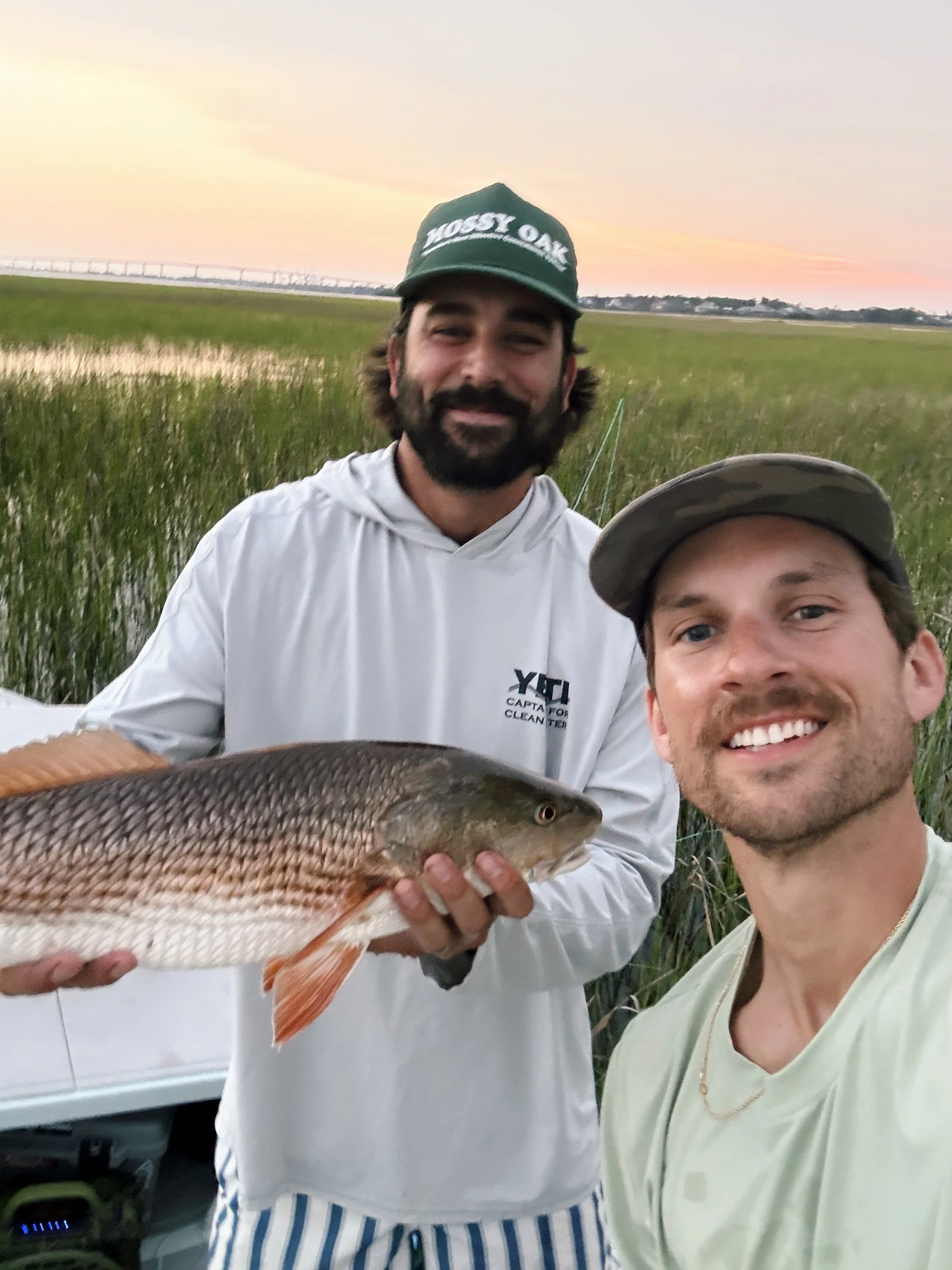 Two smiling men taking a selfie outdoors near a marsh, one holding a large fish. The man in the background wears a green hat and a white shirt, while the other wears a light green shirt and a cap. Sunset sky in the background.
