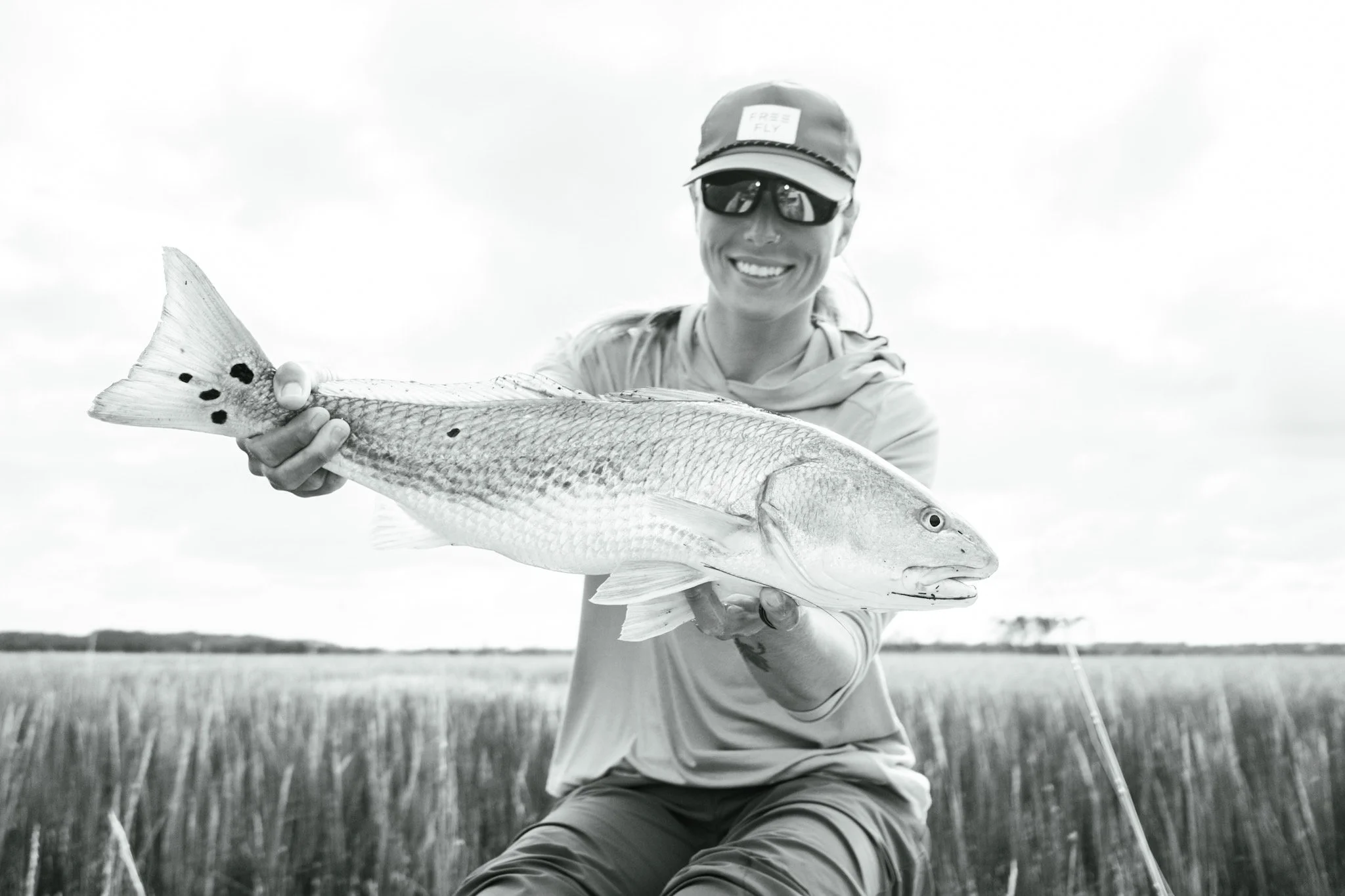 A person wearing sunglasses, a cap with a 'Free Fly' label, and casual outdoor clothing, smiling and holding a large fish in an open field.