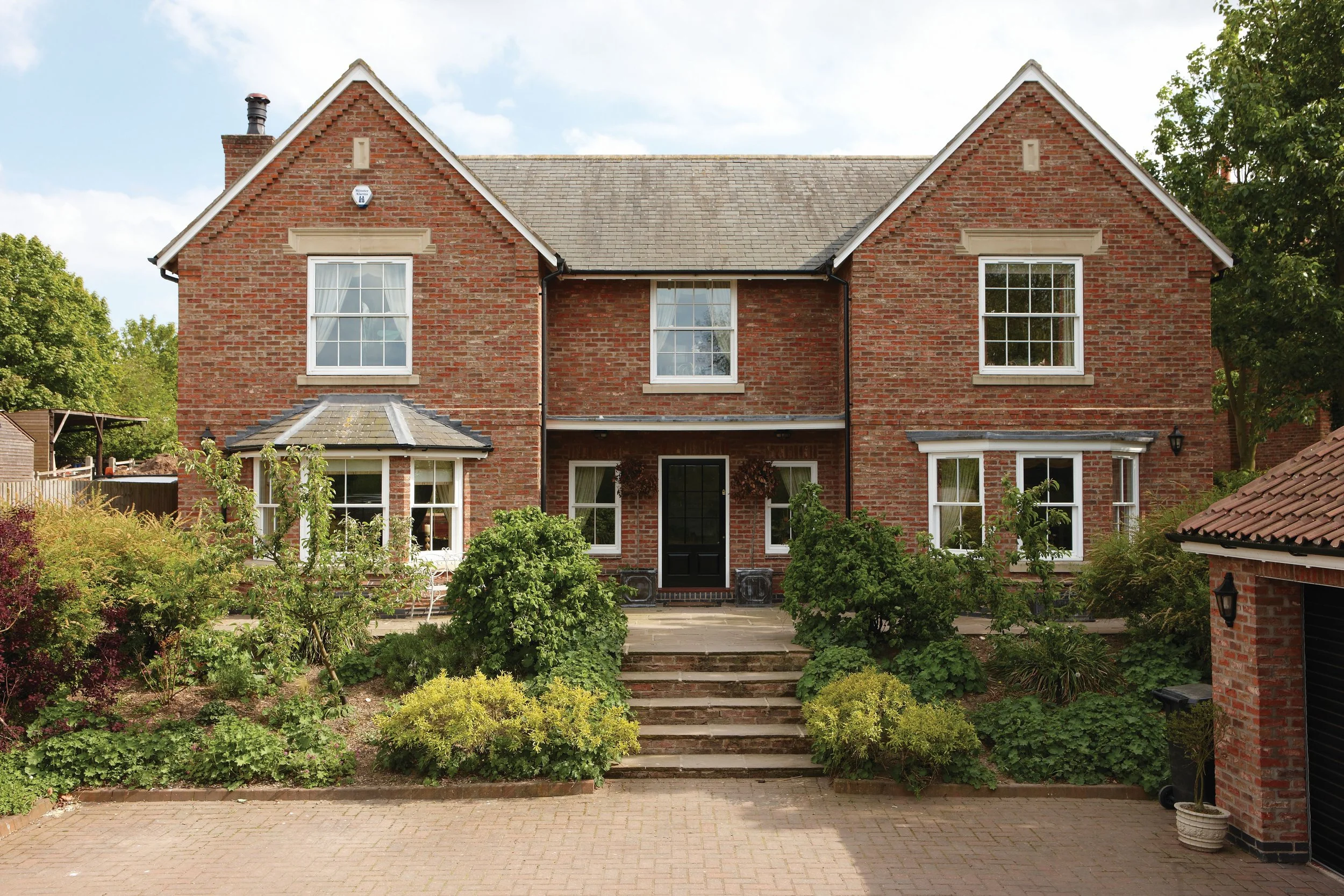 Front view of a large two-story brick house with landscaped garden and stone steps leading to black front door, surrounded by greenery and trees.