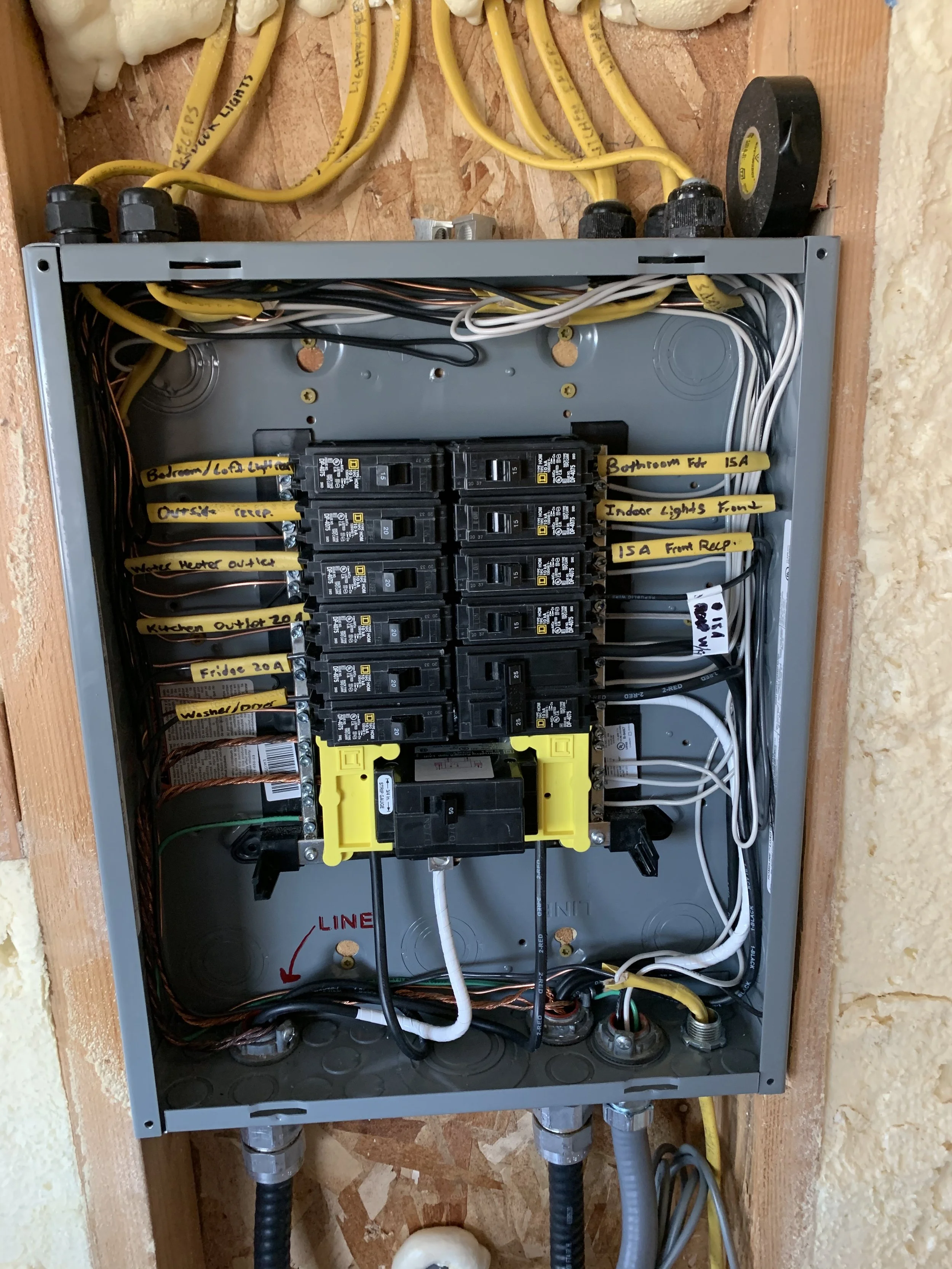 Electrical breaker box with multiple labeled yellow wires and black circuit breakers inside a metal panel, mounted on a wall with insulation around.